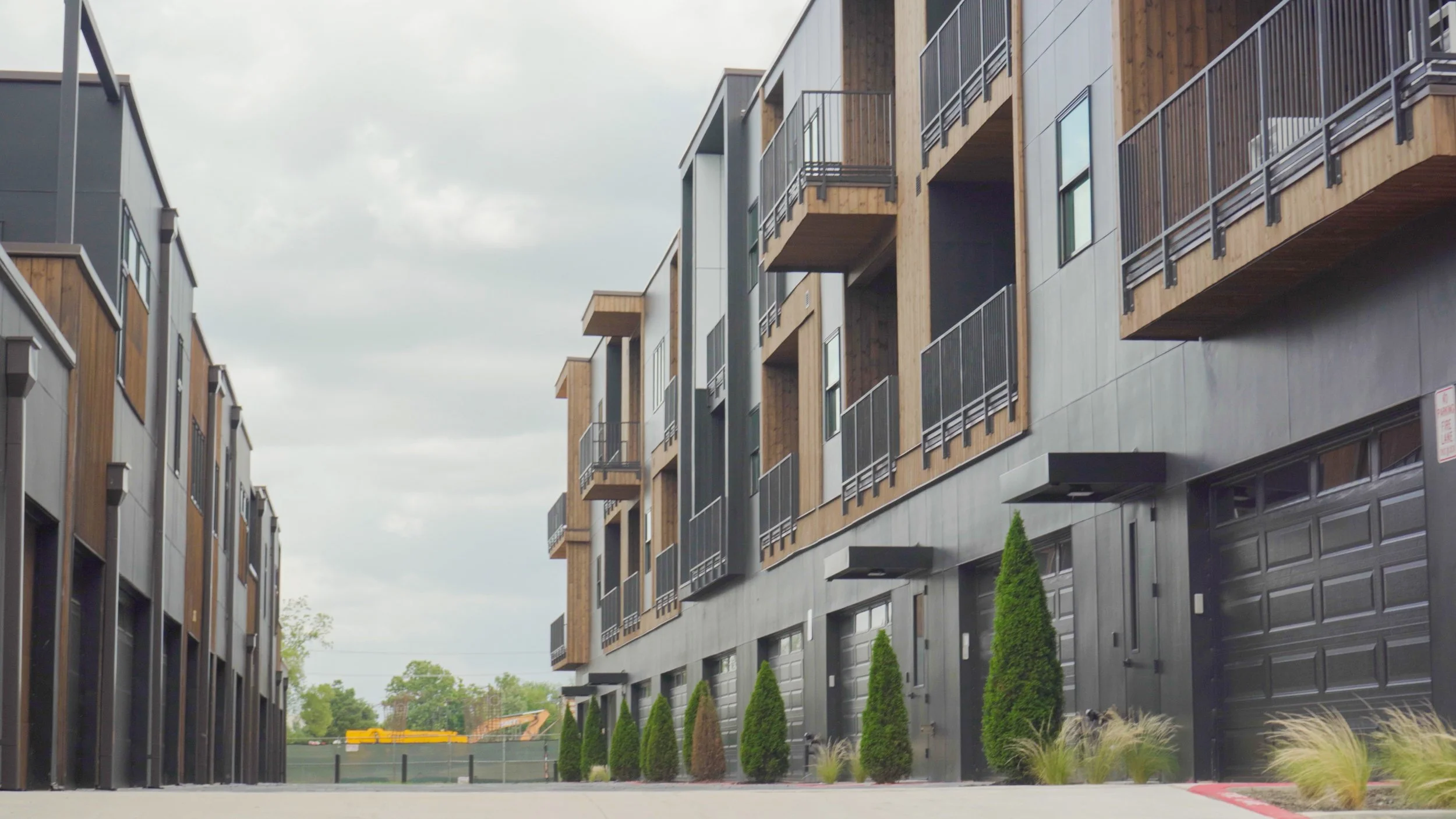 Modern multi-story residential building with a mix of wood and dark-colored exterior panels, balcony railings, and small landscaped shrubs along the sidewalk.