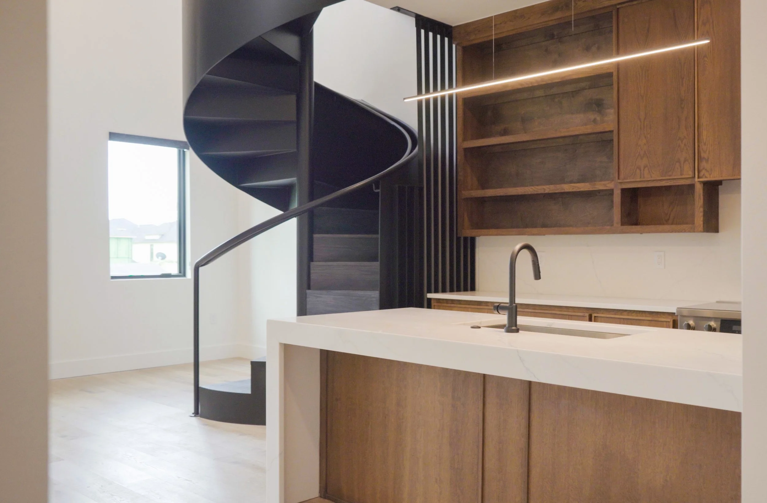 Interior kitchen with a white island, wooden cabinets, a black spiral staircase, a window, and modern lighting.