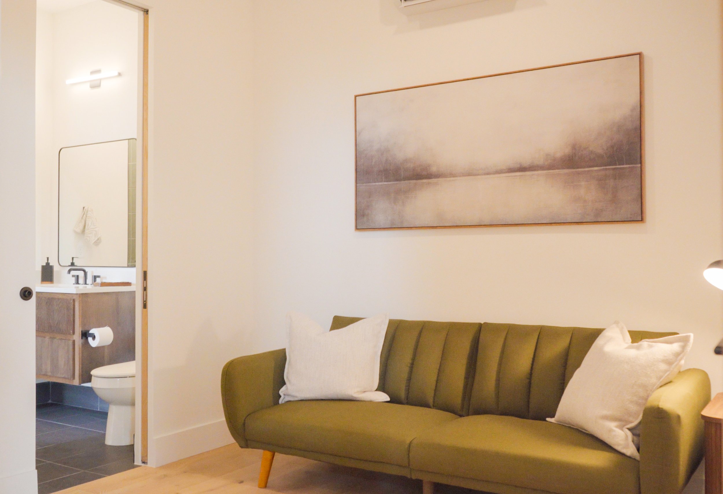 A living room with a green sofa with cushions, a wooden side table, a large abstract painting on the wall, a mirror and a bathroom with a toilet visible in the background.