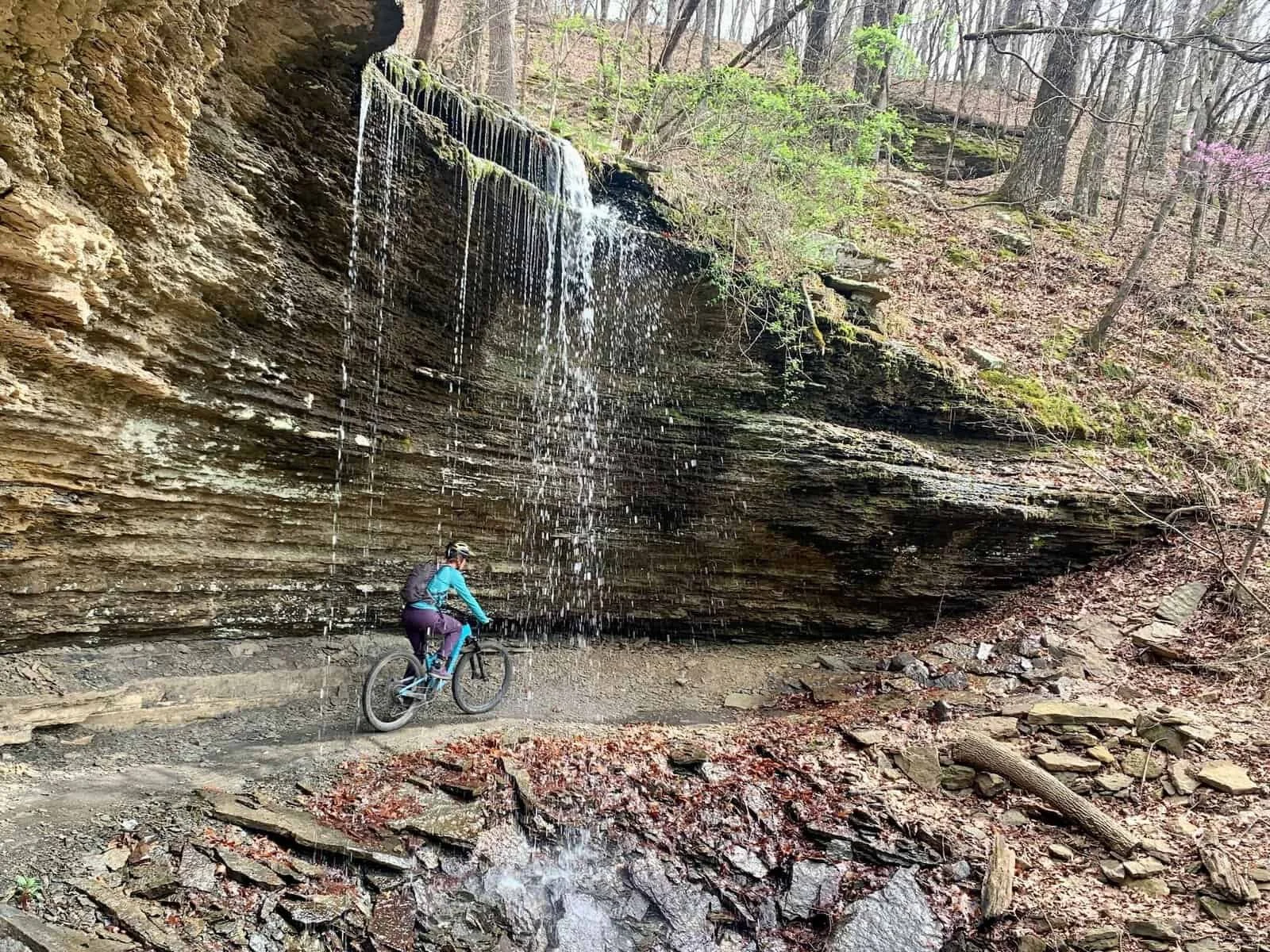A person mountain biking on a trail near a small waterfall and rock formation in a forested area during daytime.
