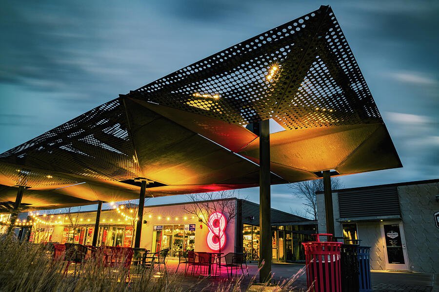 Outdoor shopping center with modern metal canopies, illuminated by warm lights at dusk, featuring a pink neon number 8 sign.