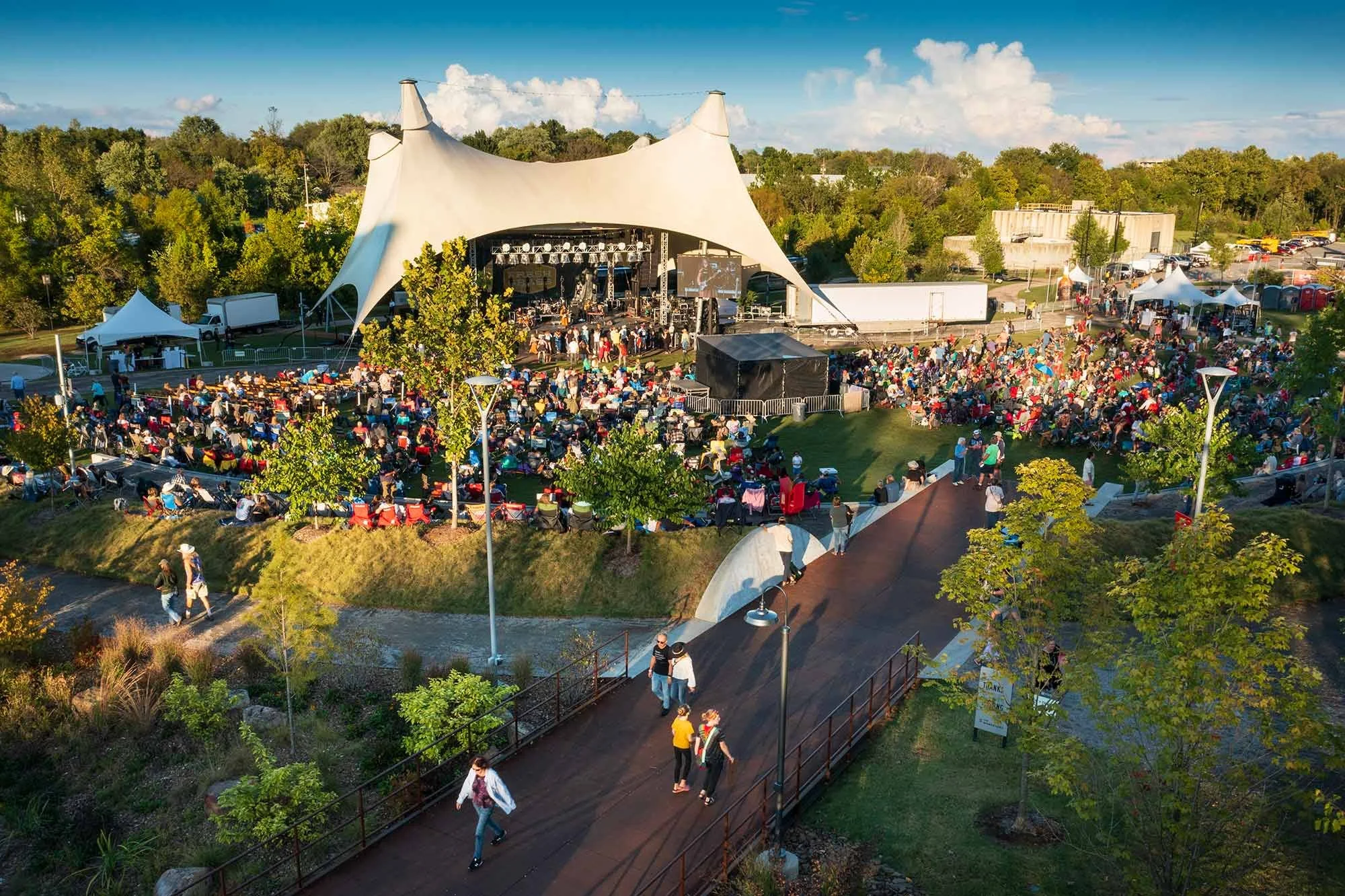 Outdoor concert at a large festival, with a stage under a white tent, a crowd seated and standing, surrounded by trees and vendor tents, on a clear day with blue sky and some clouds.