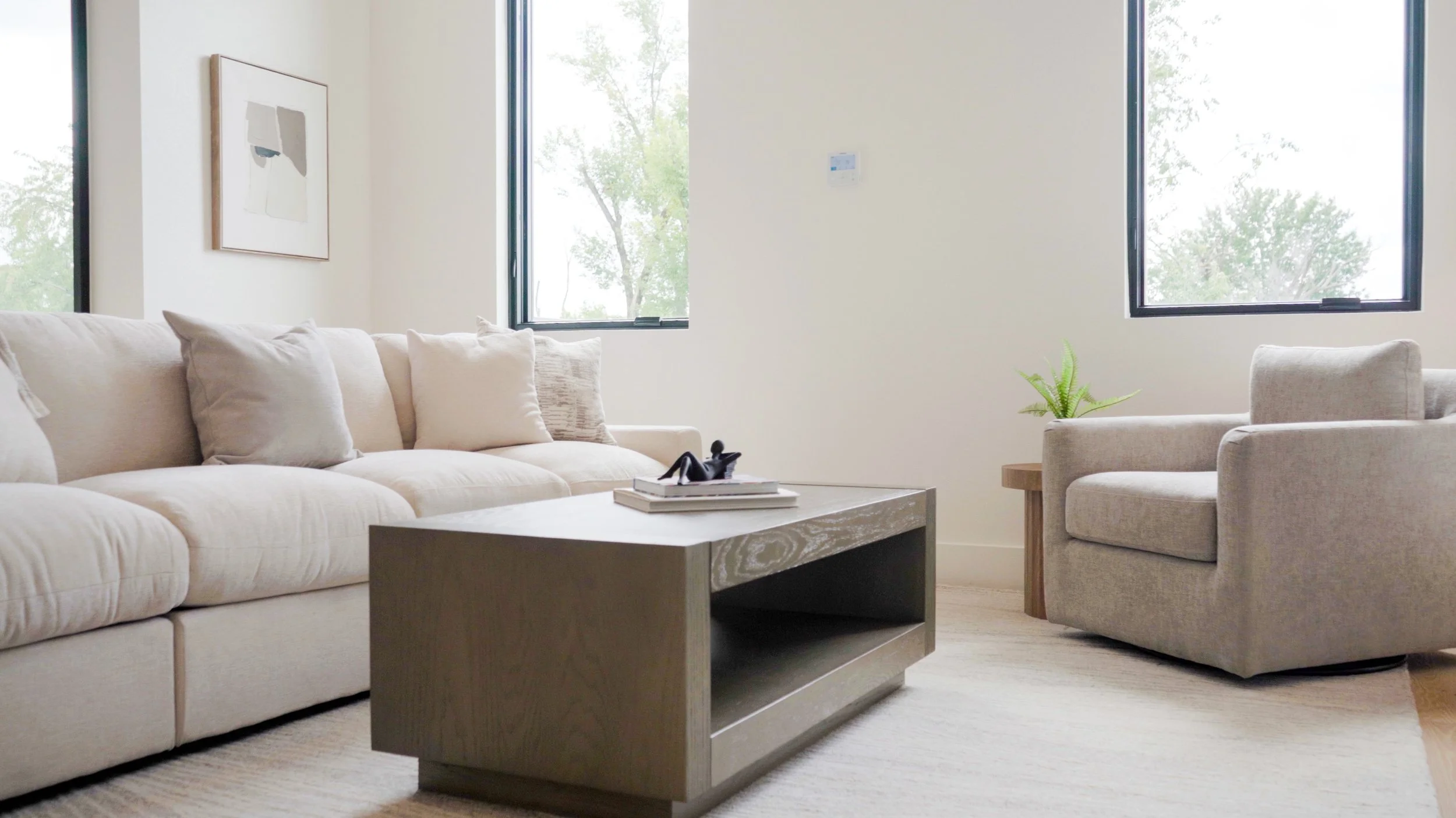 A modern living room with a cream-colored sofa and matching armchair, a light wood coffee table, and two large windows allowing natural light, with a small green plant on a side table and abstract wall art.