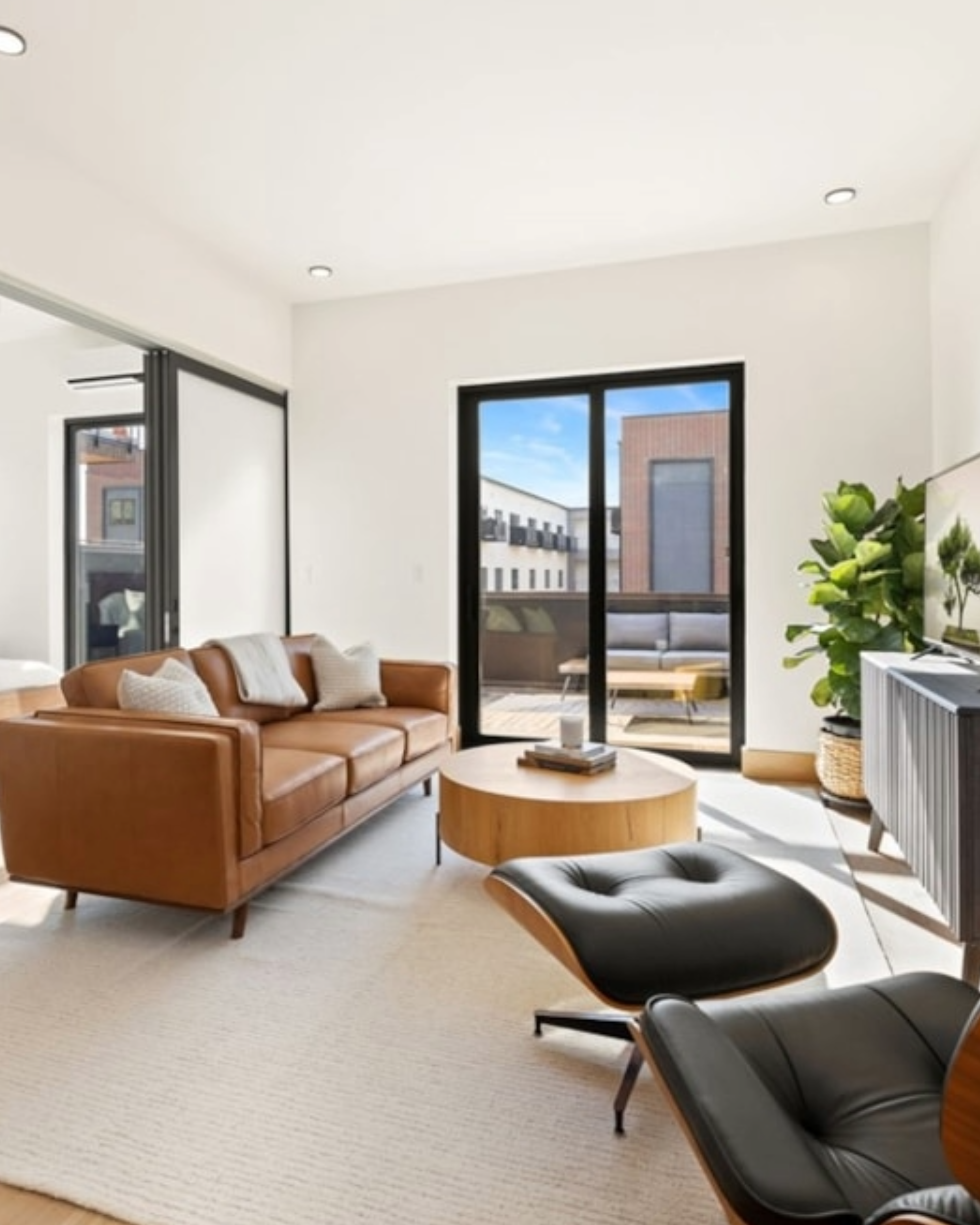 Living room with a leather sofa, modern lounge chair, coffee table, potted plant, and large windows showing an outdoor patio and buildings outside.