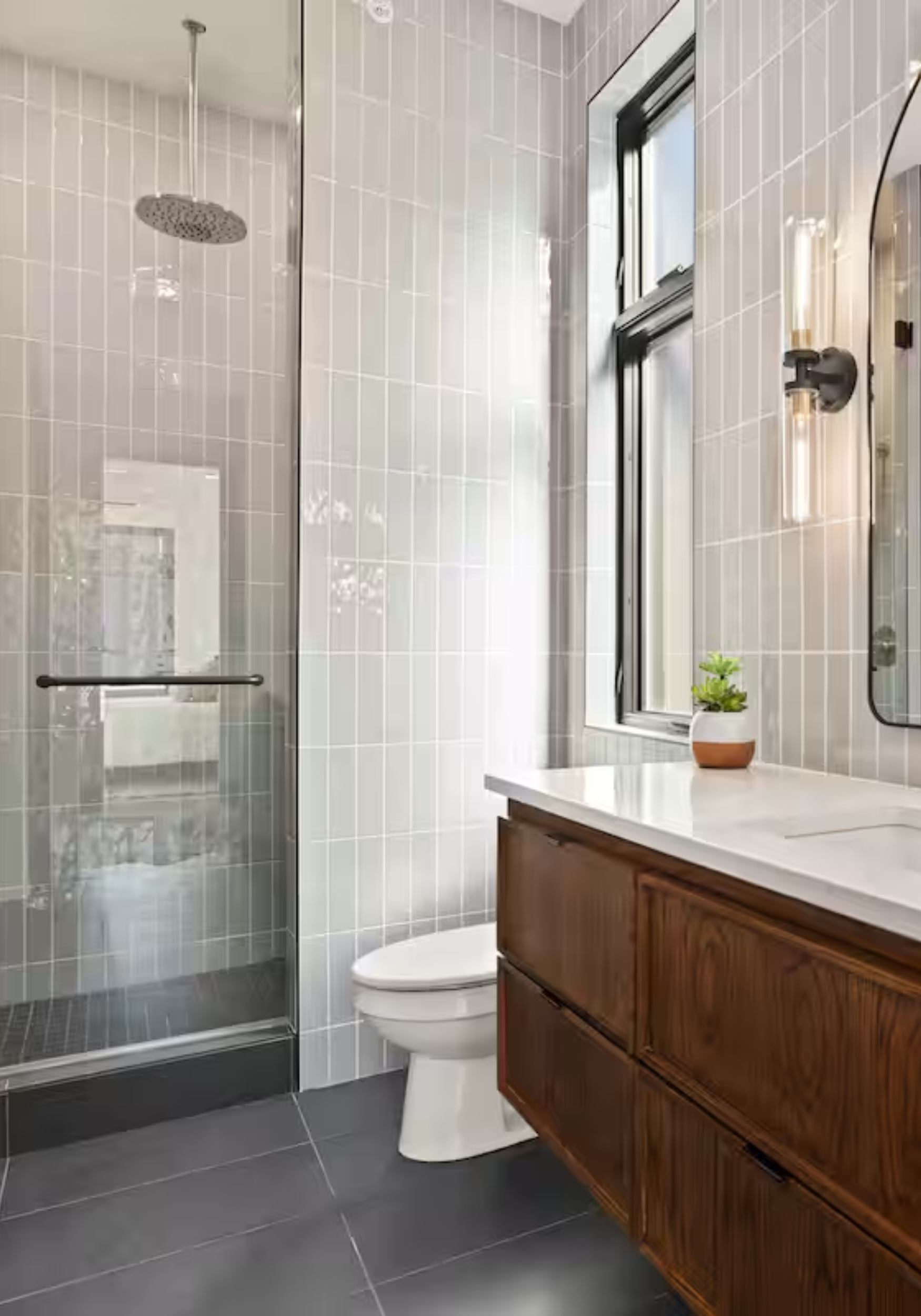 Modern bathroom with a glass shower enclosure, a white toilet, a wooden vanity with a white countertop, a window, a potted plant, and a mirror with a wall-mounted light fixture.