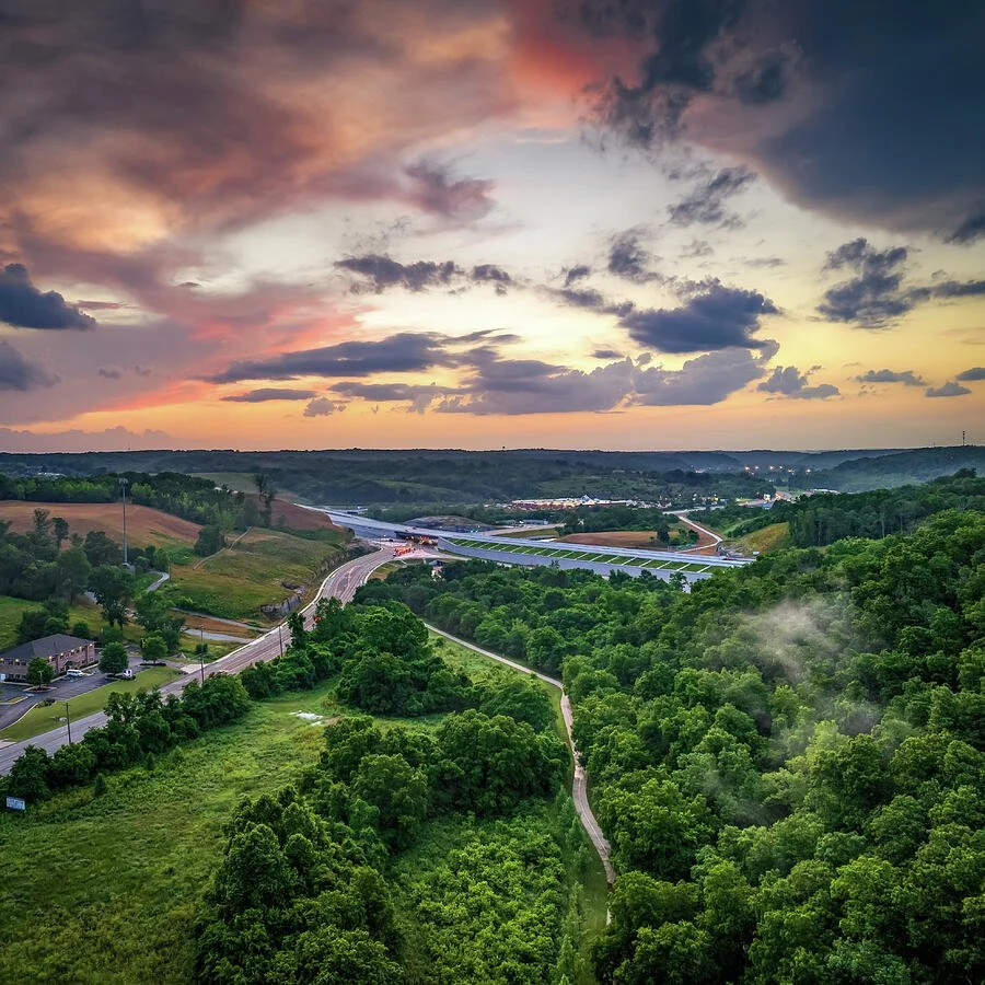Sunset over a hilly landscape with winding highways, residential buildings, and dense green forests.