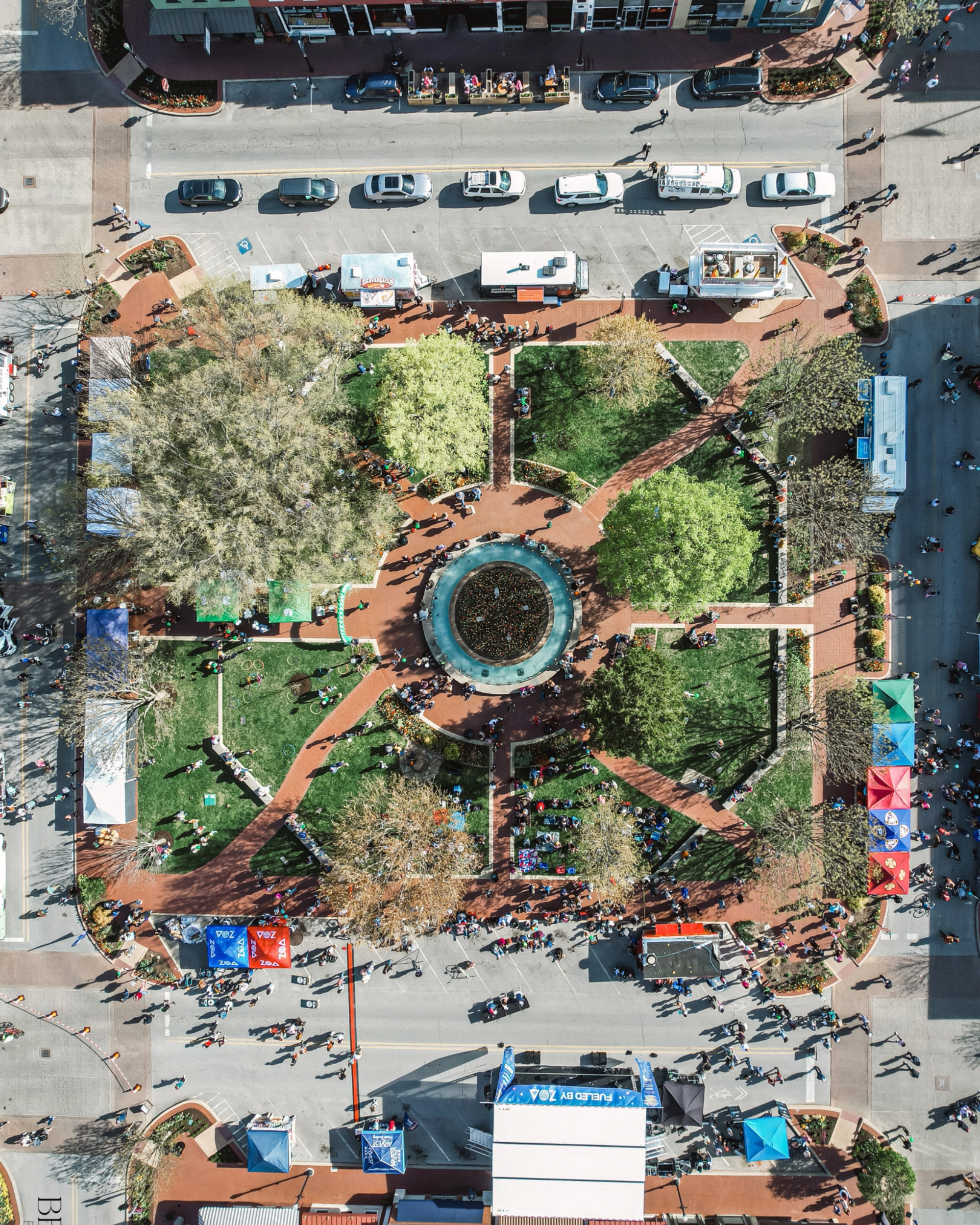 Aerial view of Downtown Bentonville with pedestrians, street vendors, trees, and tents in a park area. Parked cars line the street at the top of the image.