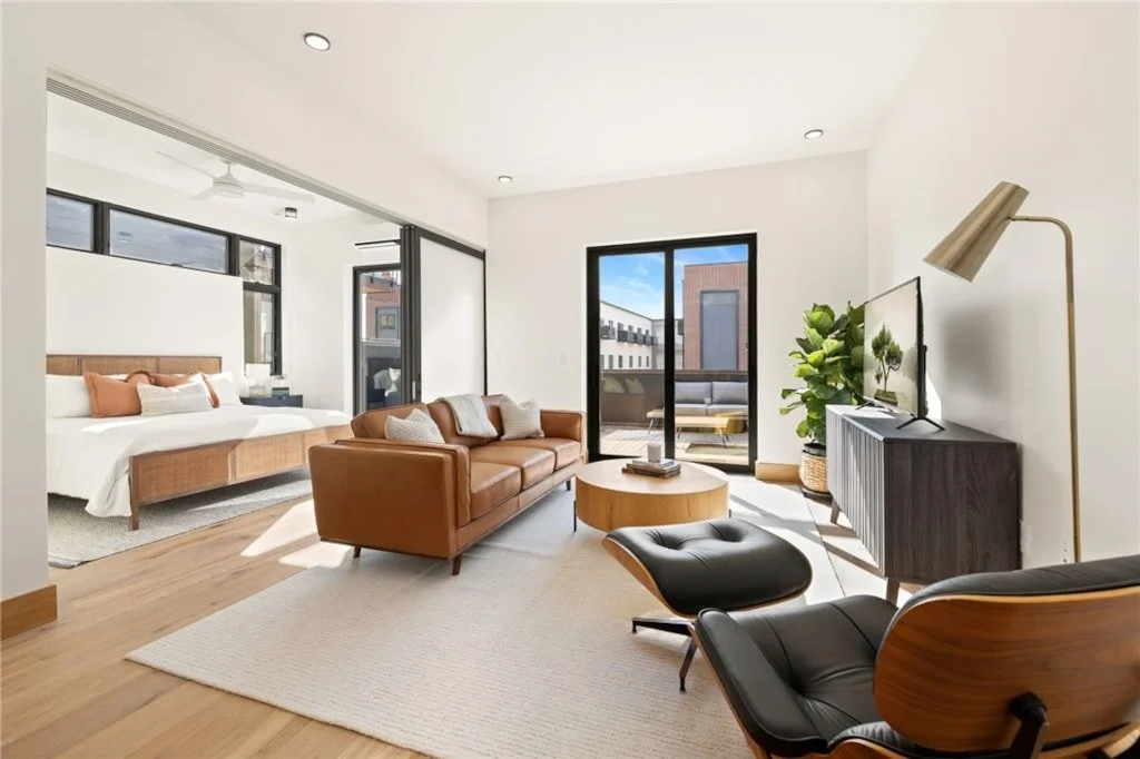 Bright living room with a brown leather sofa, black lounge chair with ottoman, TV on a dark console table, potted plant, sliding glass door leading to a balcony, and a bedroom with a bed and pillows visible through an open wall.