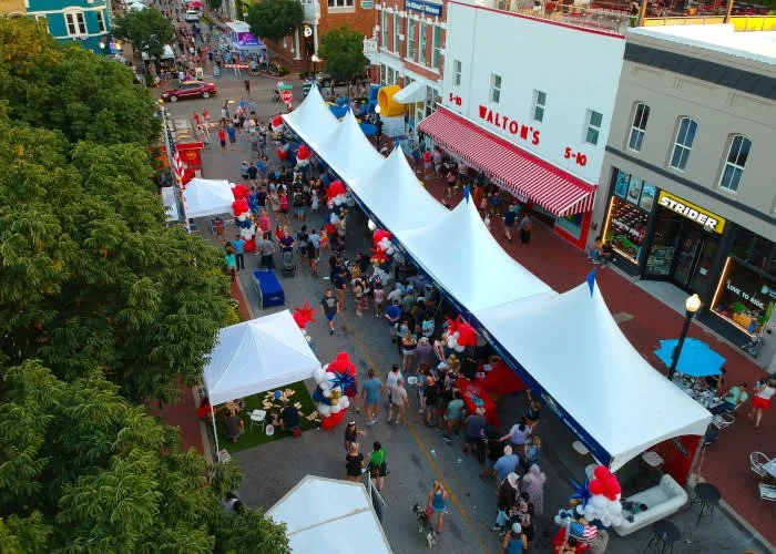 Crowd of people walking along a street decorated for an event, with white tents and balloons, and shops in the background.