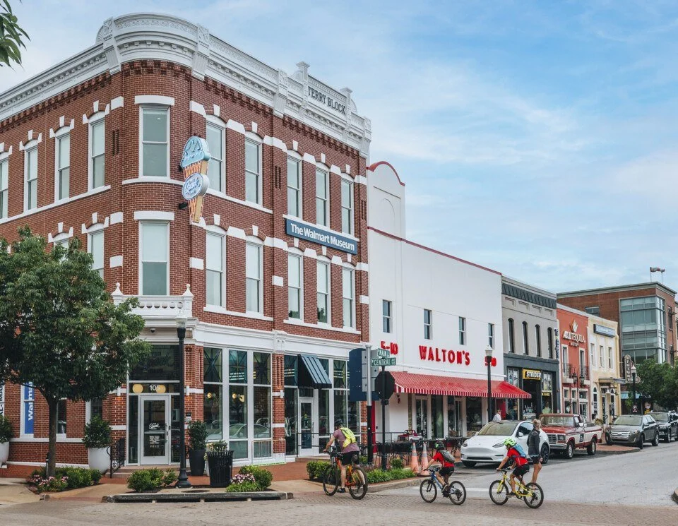 Street view featuring a brick building with a sign for The Walmart Museum, adjacent stores, parked cars, and people riding bikes.