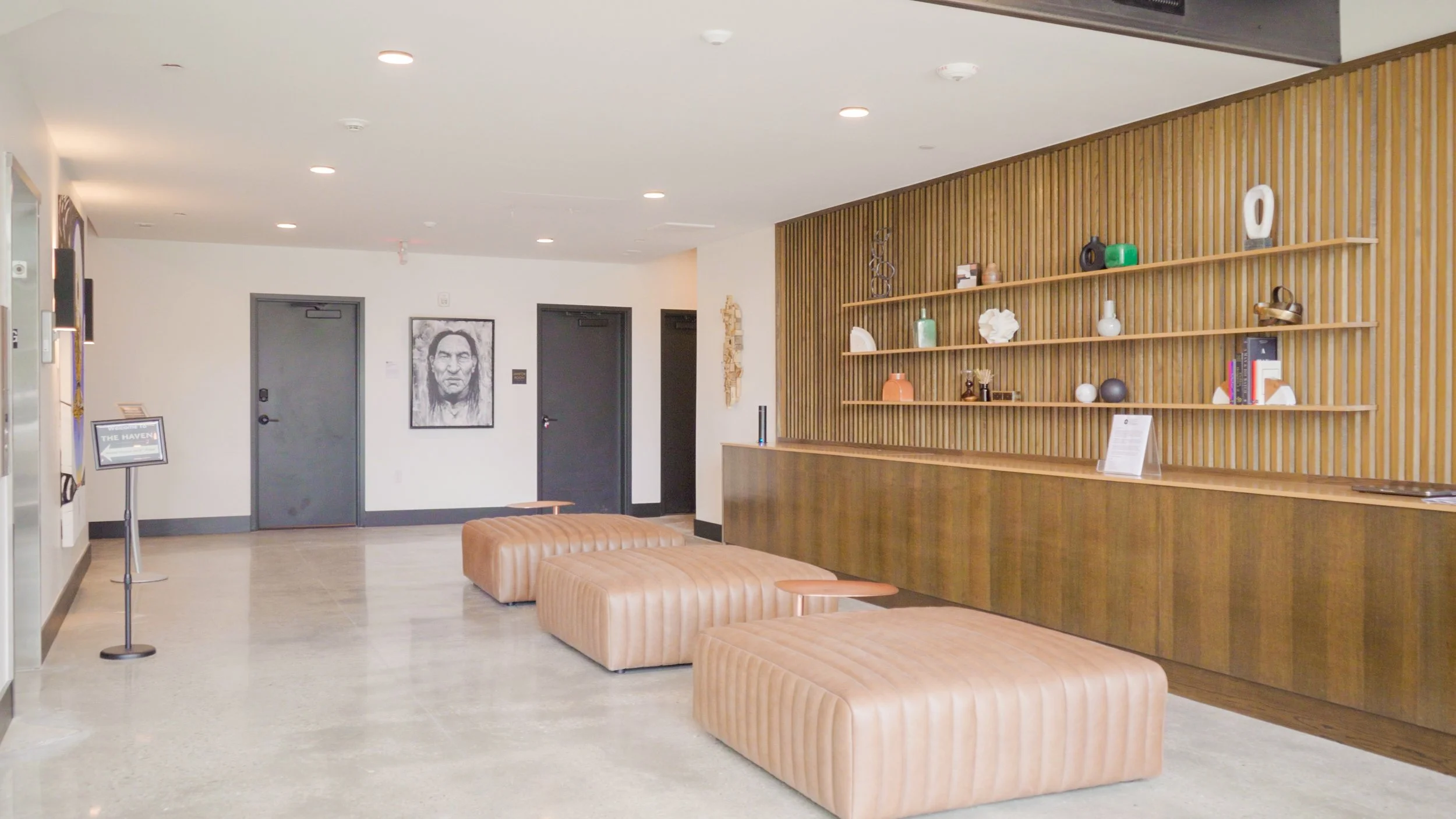 Modern area with tan leather ottomans, a wooden wall with decorative items on shelves, and black doors with framed artwork on white walls.