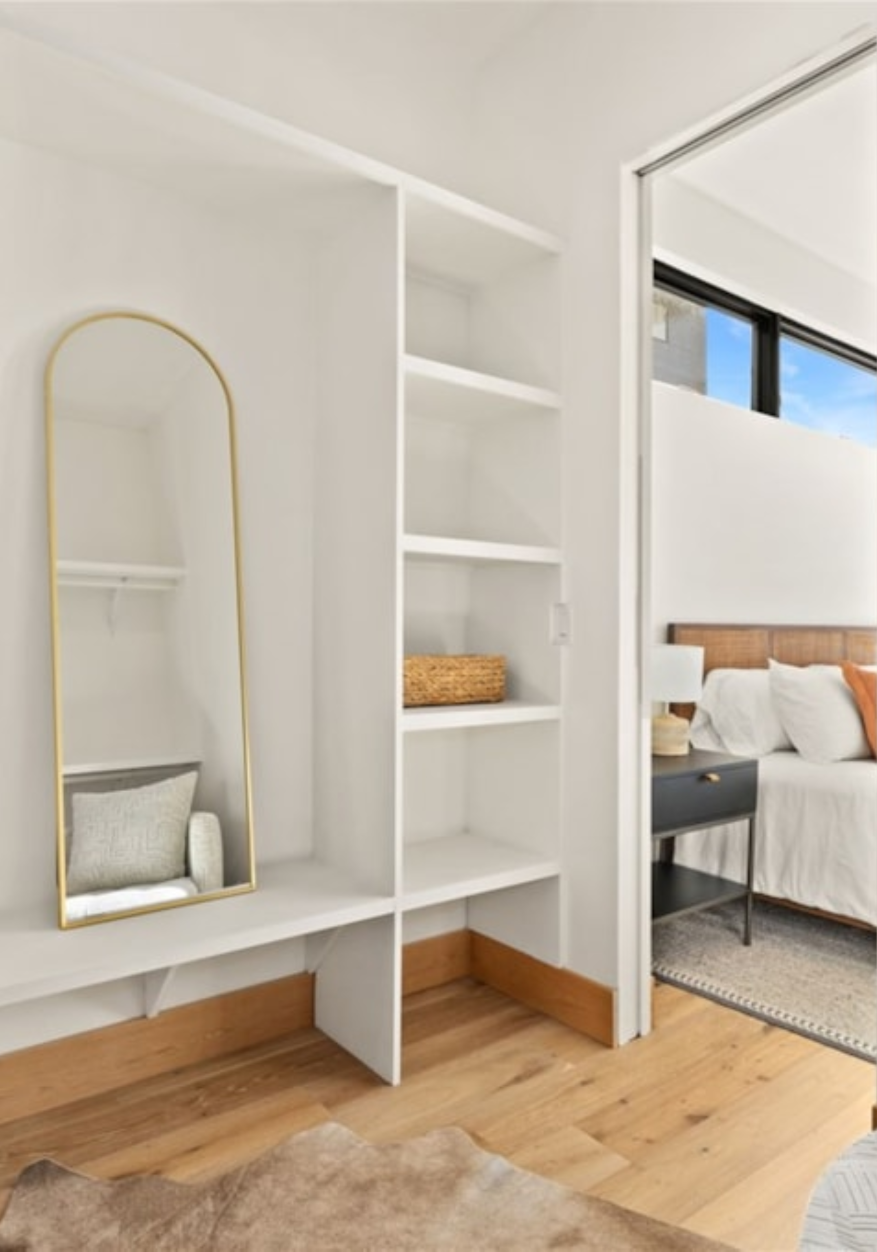 A white shelving unit with open shelves, a tall mirror with a yellow frame, and a woven basket. There is a bed with a white bedspread and orange pillows in the background, near a large window with a black frame showing a blue sky and part of a building.