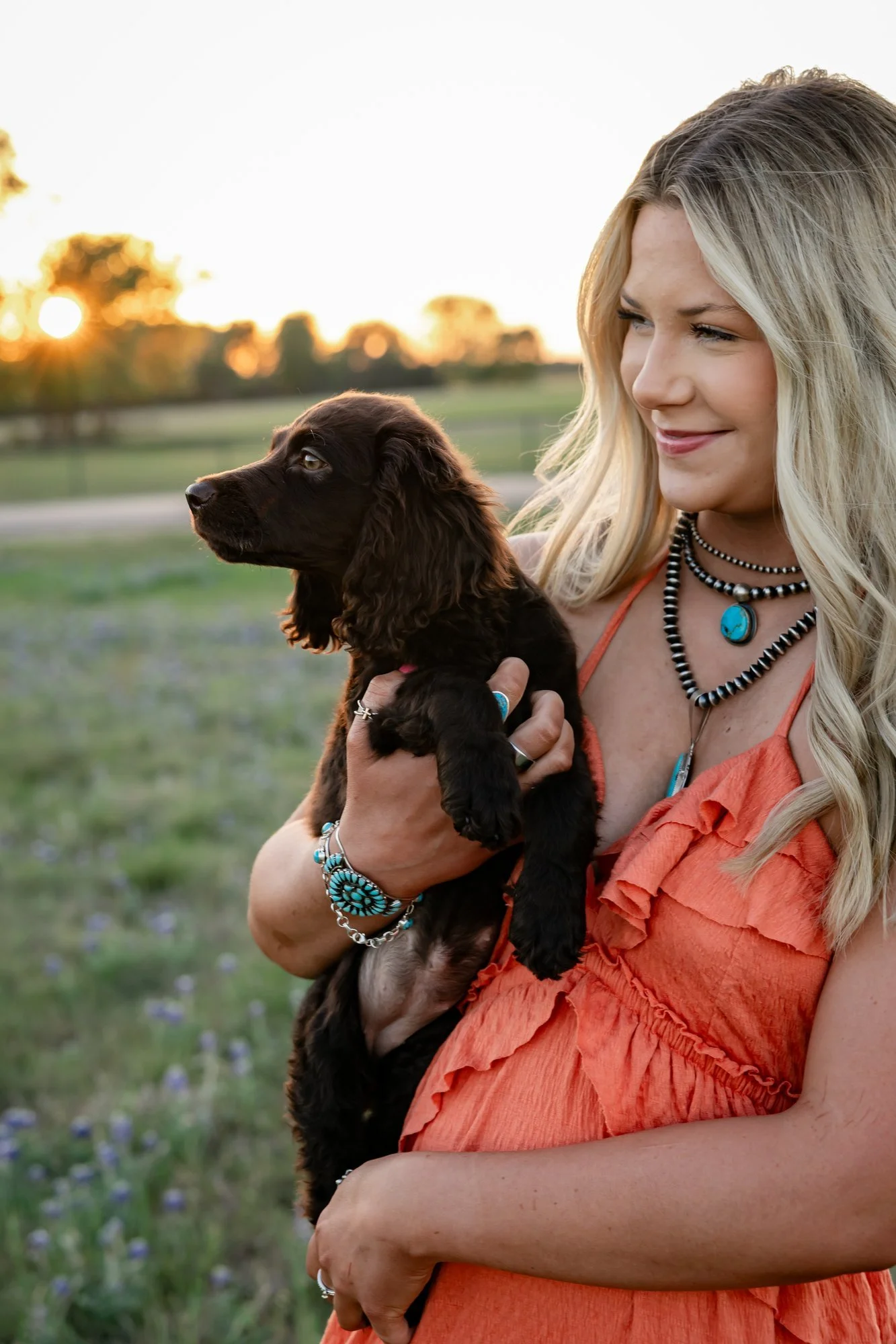 A woman with long blonde hair wearing an orange dress and turquoise jewelry holding a black puppy with brown fur, with a sunset and trees in the background.