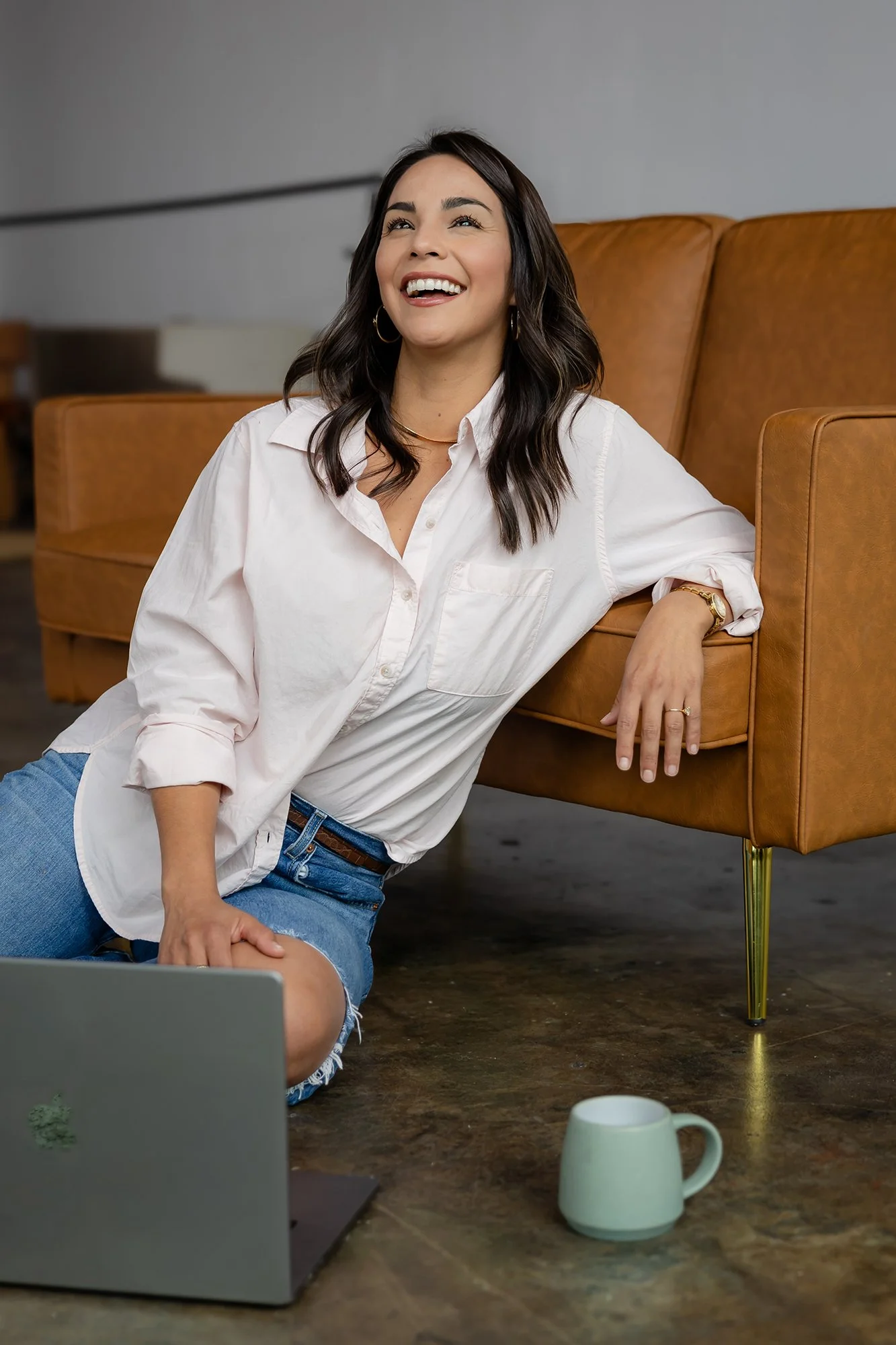 A woman sitting on the floor of Sunroom Studio in downtown Waco near a laptop with a smile, wearing a white shirt and denim shorts, with a coffee mug on a wooden table in front of her.