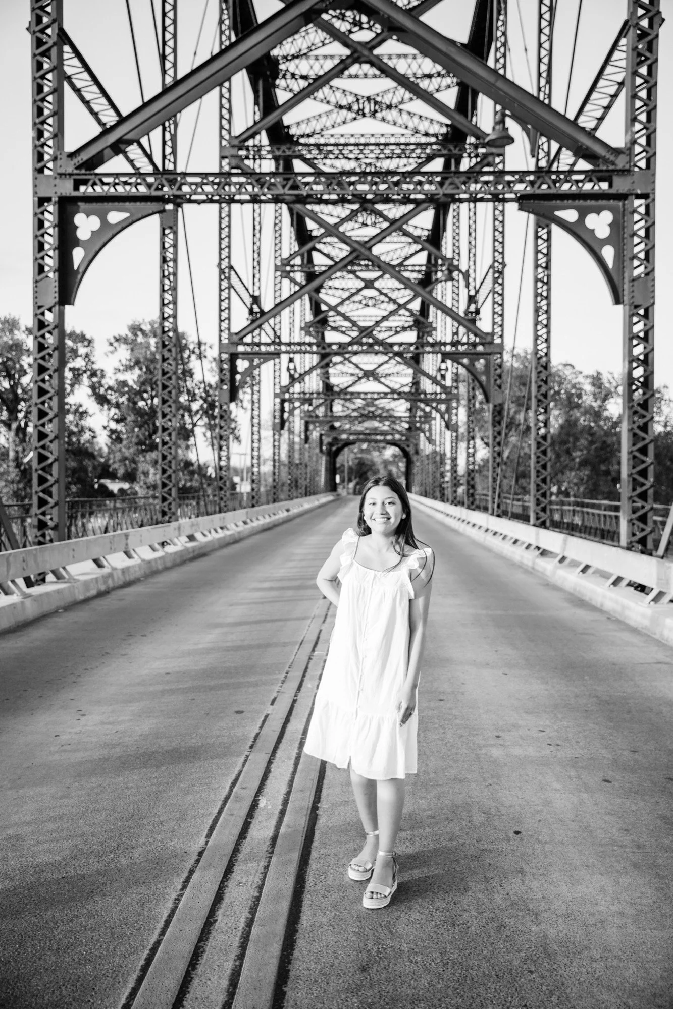 Baylor University senior standing near the Washington Avenue Bridge, a black steel truss bridge over the Brazos River in Waco, Texas.