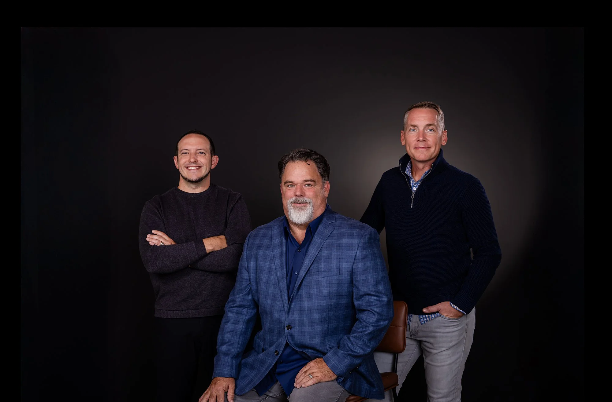 Four men posing for a photo in a professional studio. The man in front is seated on a chair, wearing a blue plaid blazer, and the other three are standing behind him, smiling. The background is black.