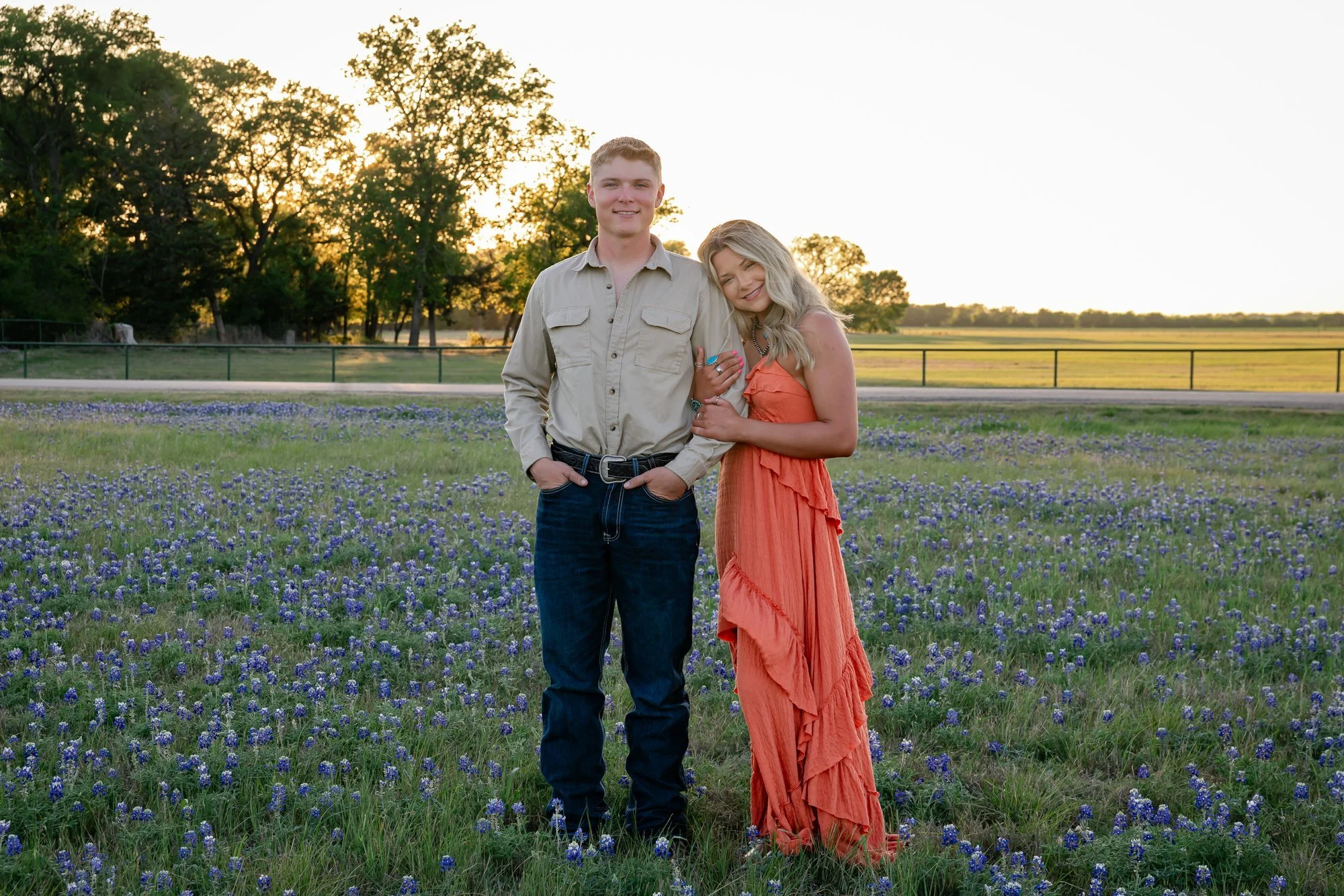 A young man and woman standing close together in a field of purple flowers at sunset. The man is wearing a beige shirt and jeans, with his hands in his pockets. The woman is wearing an orange dress, smiling and leaning on the man's shoulder, holding his arm. There are trees and open fields in the background.