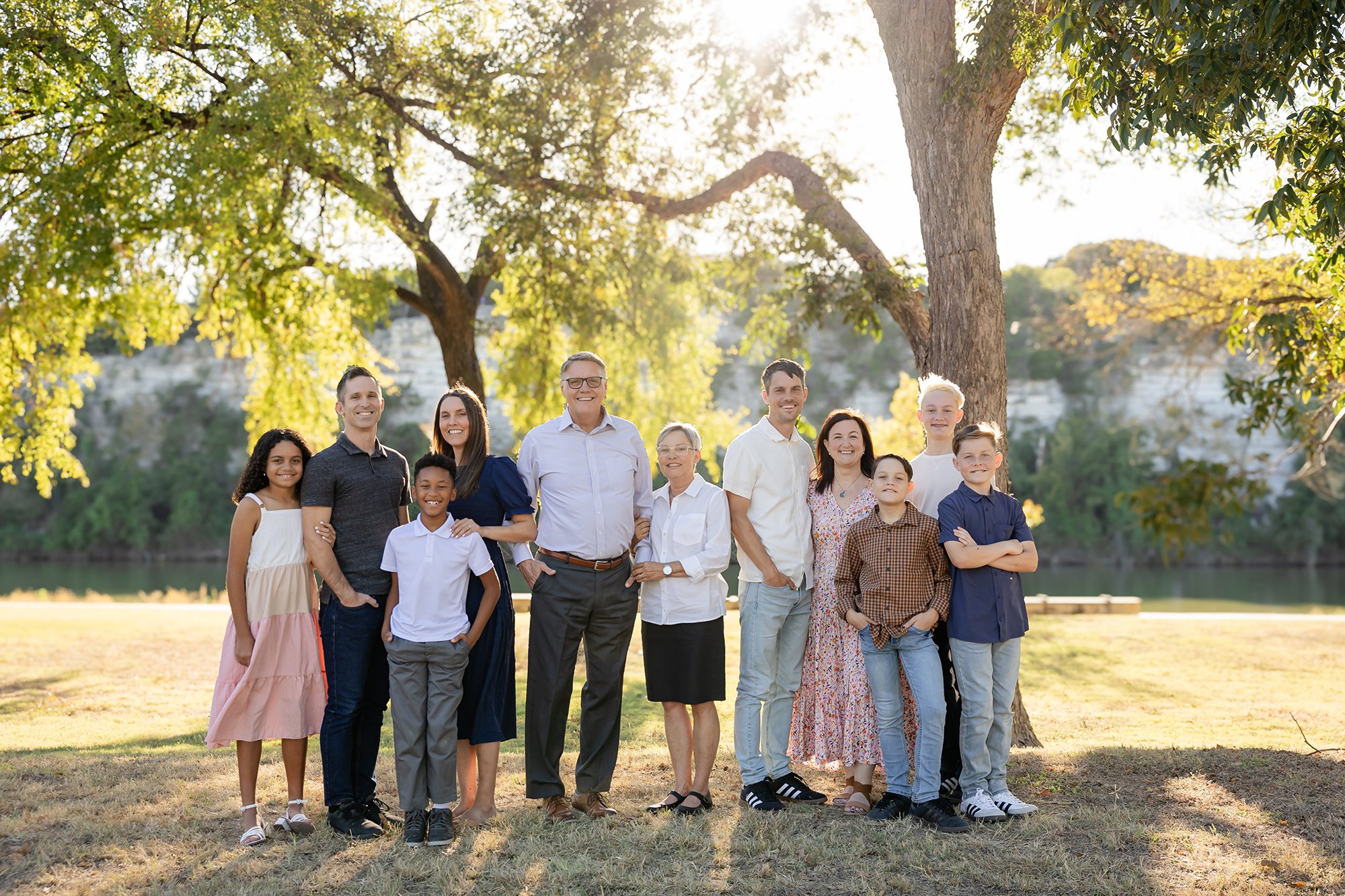 A diverse group of people, including children, adults, and seniors, standing and smiling outdoors in a park with trees and sunlight in the background.