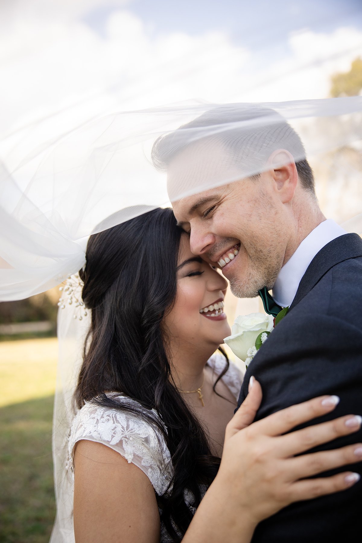 Bride and groom smiling beneath the bride’s flowing veil during an outdoor wedding portrait.