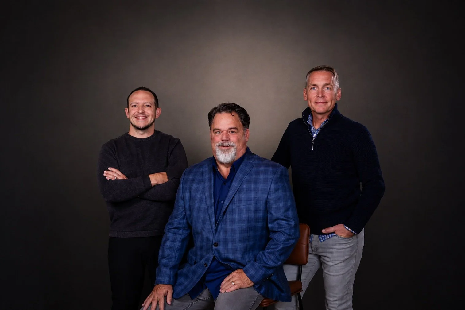 Three men posing for a group photo against a dark background. The man in the front is seated and wearing a blue plaid blazer, the man on the left has arms crossed and is wearing a dark sweater, and the man on the right has one hand in his pocket and is wearing a dark jacket over a checkered shirt.