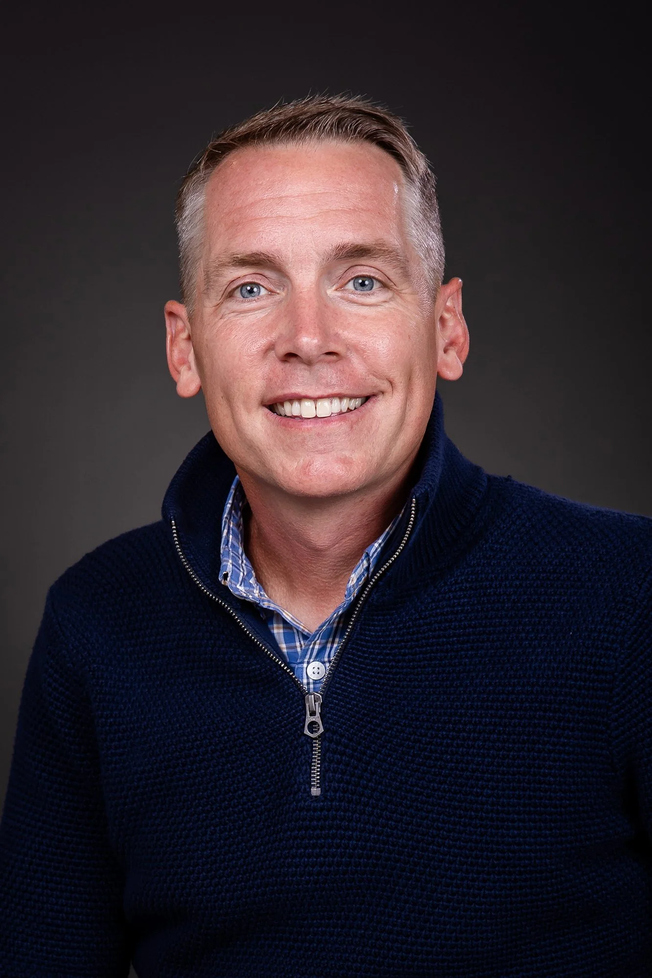 Headshot of a smiling man, Clint Harp of Harp Design Co, with blue eyes, short light brown hair, wearing a dark blue knitted zip-up sweater over a plaid button-up shirt against a dark background.