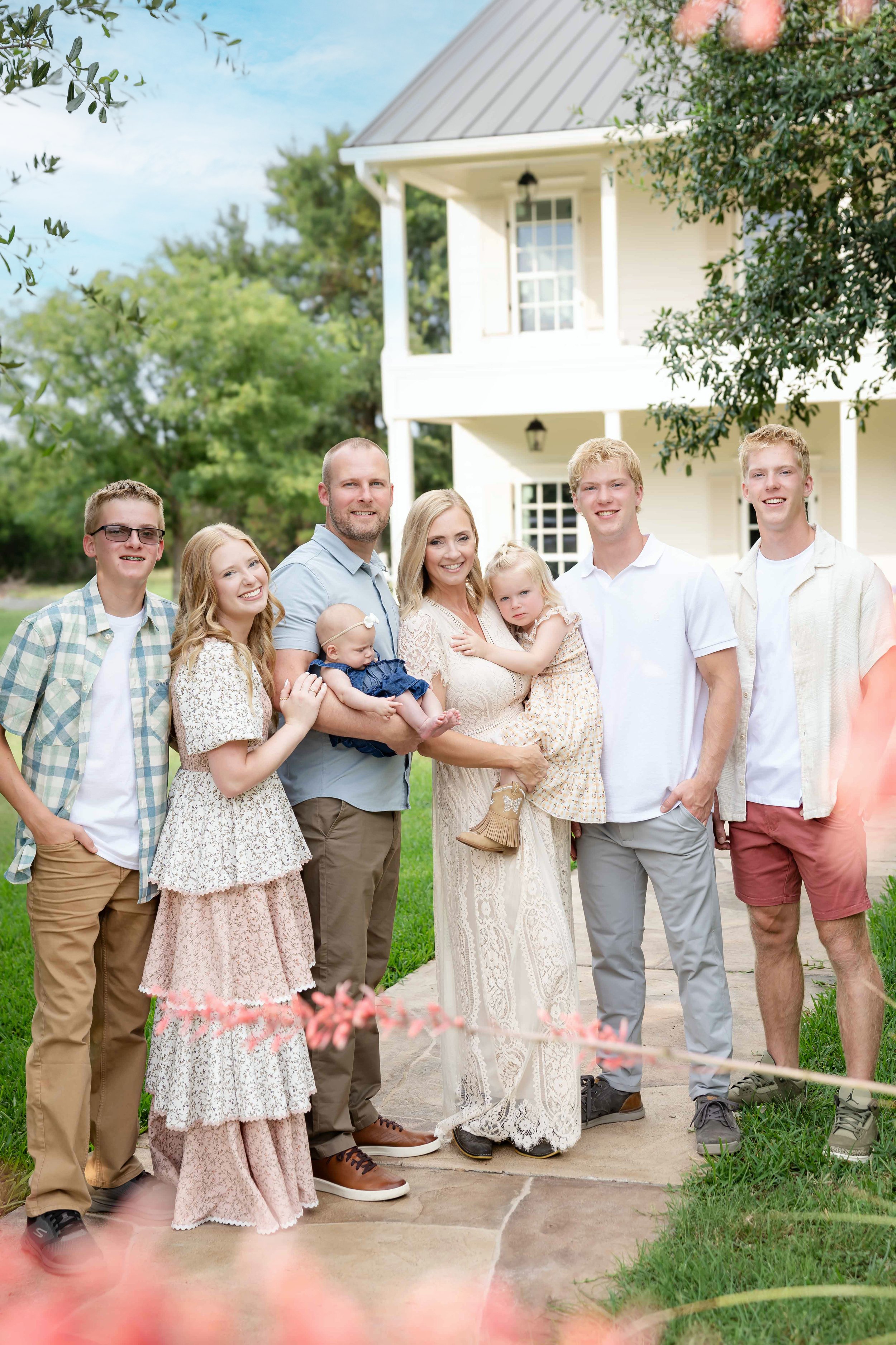 Family photo of eight people standing outside in front of a white house with a balcony, greenery, and pink flowers in the foreground. The group includes adults and children smiling at the camera.