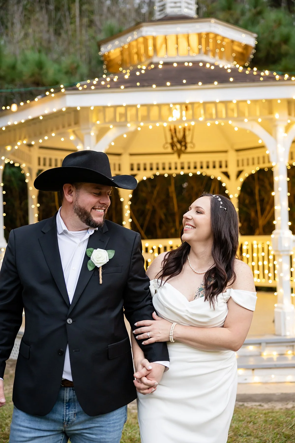 A couple at a wedding or special event, standing outdoors in front of a lit gazebo at dusk. The man is wearing a black cowboy hat, a black blazer with a white shirt, and jeans, with a white rose boutonniere. The woman is in a white off-the-shoulder dress with pearl jewelry and a hair accessory. They are smiling at each other and holding hands.