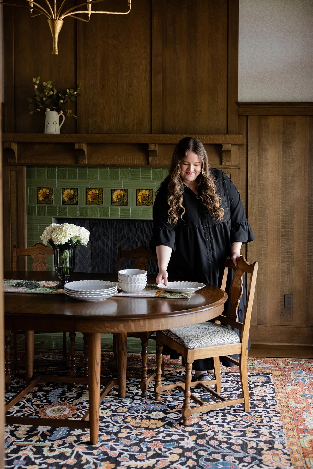 A woman in a black dress is setting a dining table with white plates and bowls, a vase of white flowers, in a room with wooden paneling, a fireplace with green tiles, and a patterned rug.