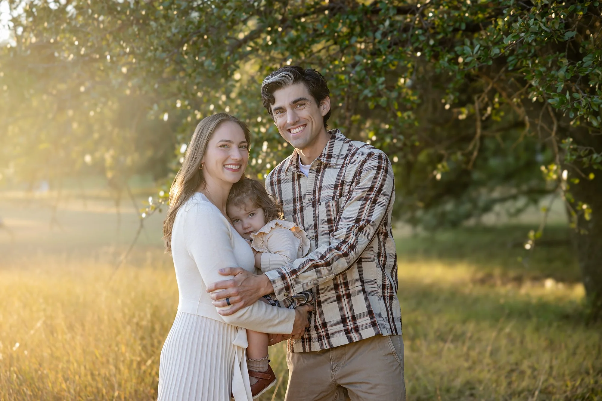 A happy family of three, a woman, a man, and a young girl, standing outdoors in a sunny park with green trees in the background. The woman has long blonde hair and the man has dark hair. The girl has curly hair and is being held by the woman.