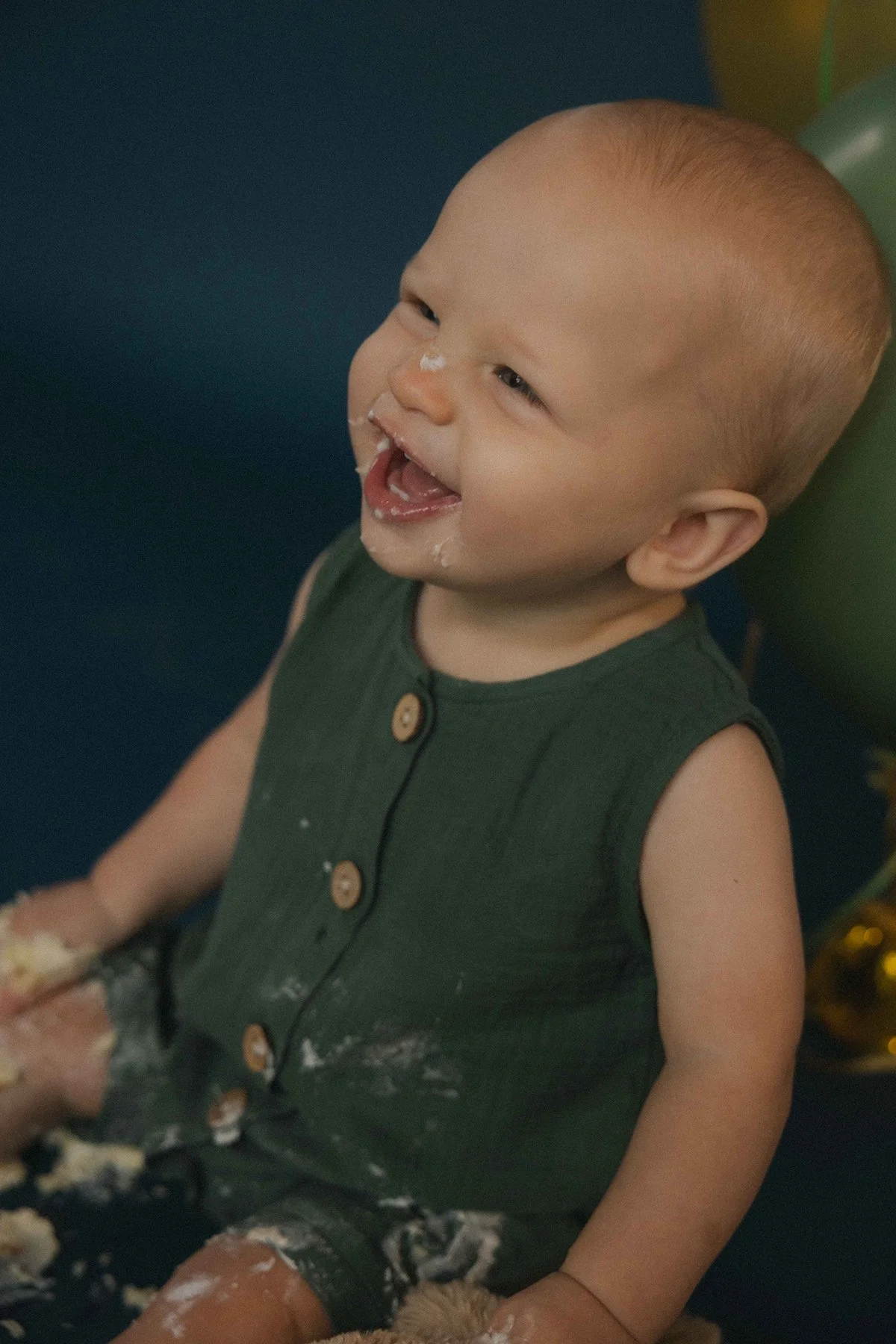 Smiling baby with short hair, playing with cake and having white frosting on face, wearing a sleeveless dark green shirt with wooden buttons.