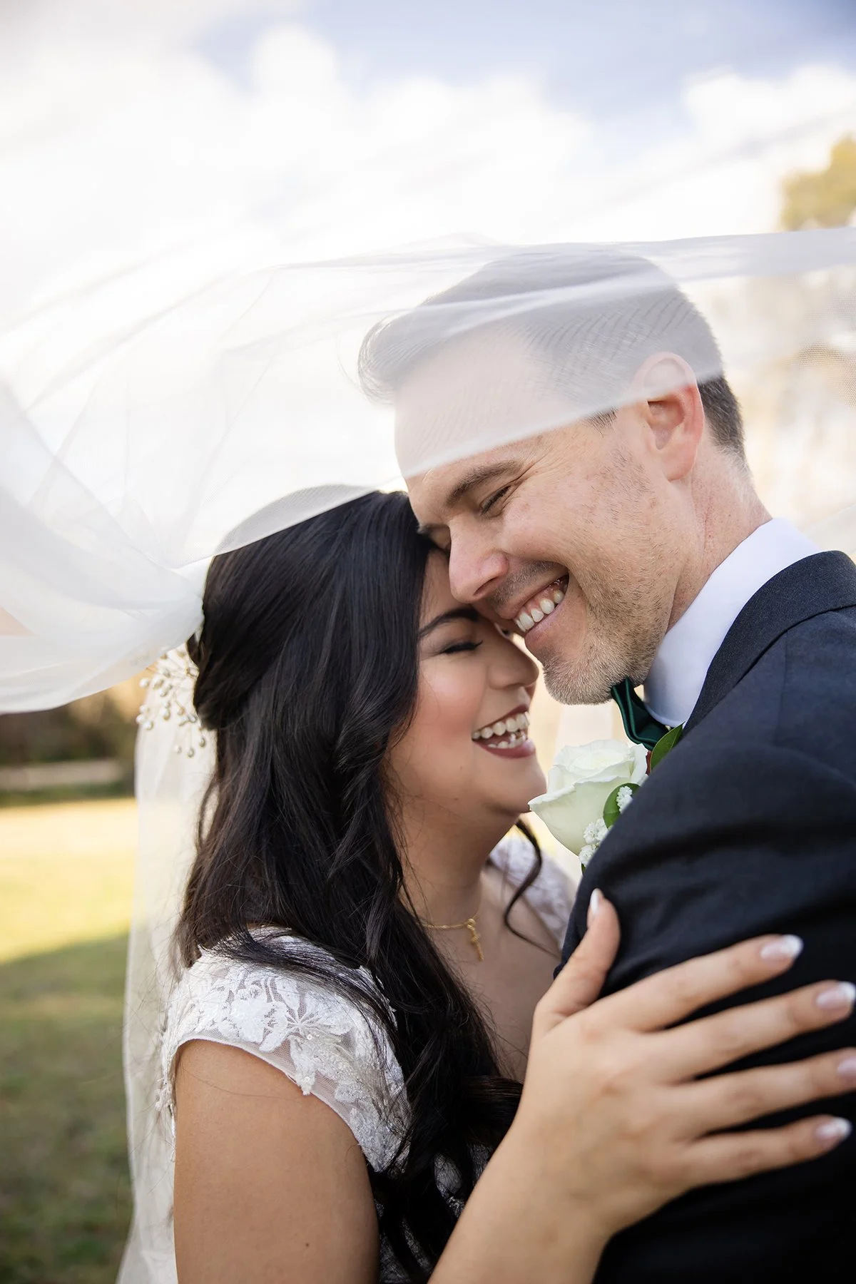 A happy bride and groom embracing outdoors, close-up of their faces under a veil, smiling and enjoying their wedding moment.