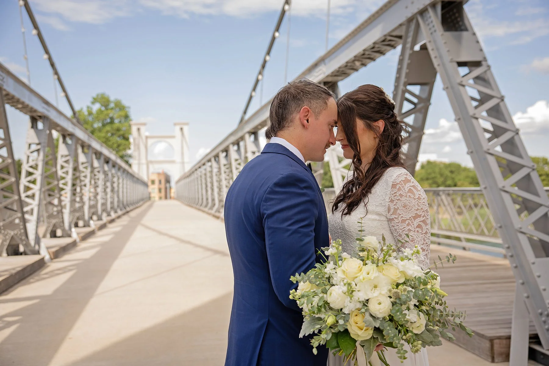 A bride and groom standing close on a bridge, touching foreheads, with the bride holding a large bouquet of white and green flowers.