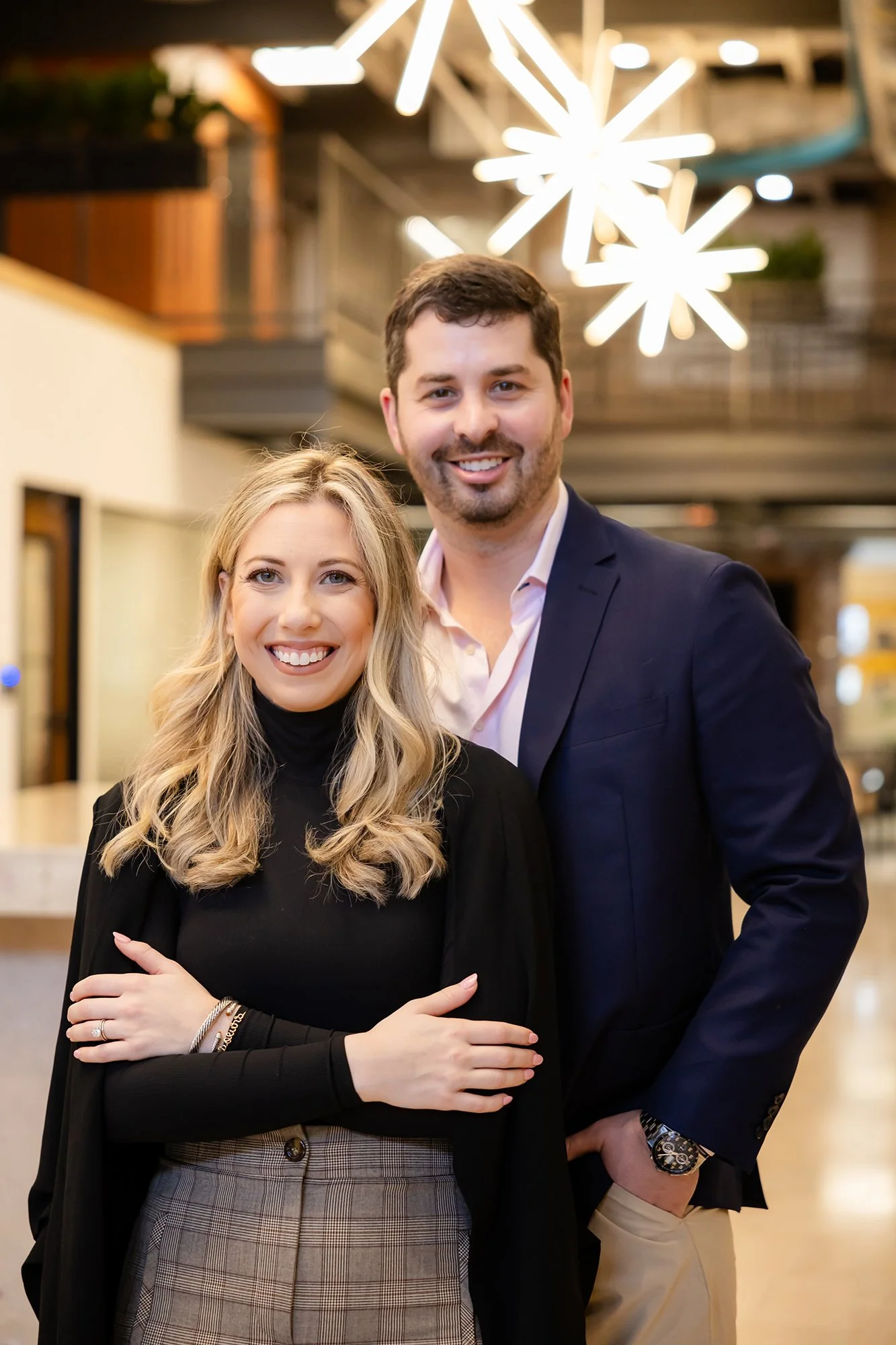 A smiling woman with long blonde hair and a man with dark hair and a beard posing together in a well-lit, modern indoor space with contemporary lighting fixtures.