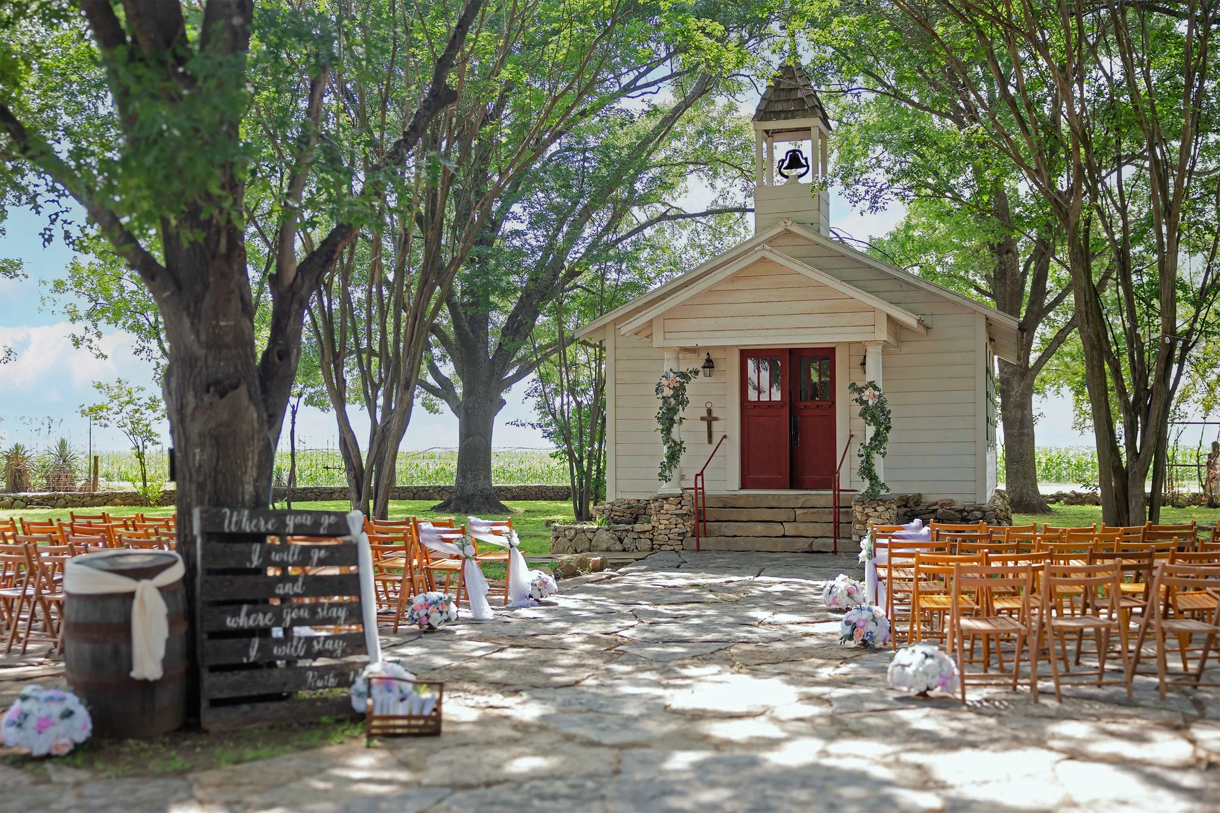 A small white chapel with a bell tower, set in a green, leafy outdoor area, decorated for a wedding with floral arrangements and chairs arranged for an outdoor ceremony.