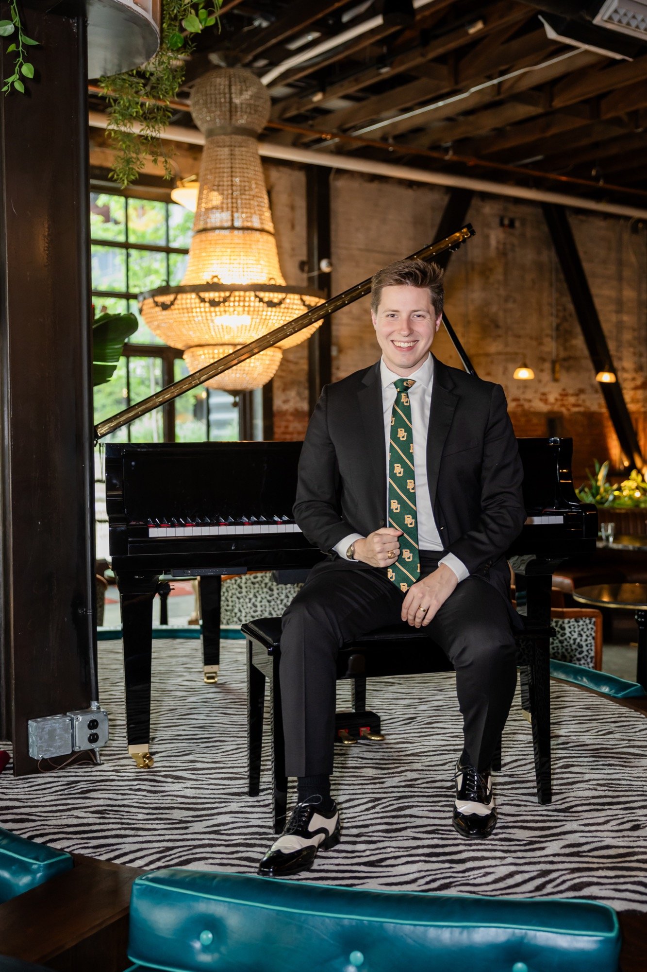 A young man dressed in a black suit with a green tie and matching pocket square, sitting on a black piano bench in a stylish venue, smiling at the camera. Behind him is a black grand piano, an ornate chandelier, and a rustic exposed brick wall. The area has large industrial-style windows, some plants, and warm lighting.
