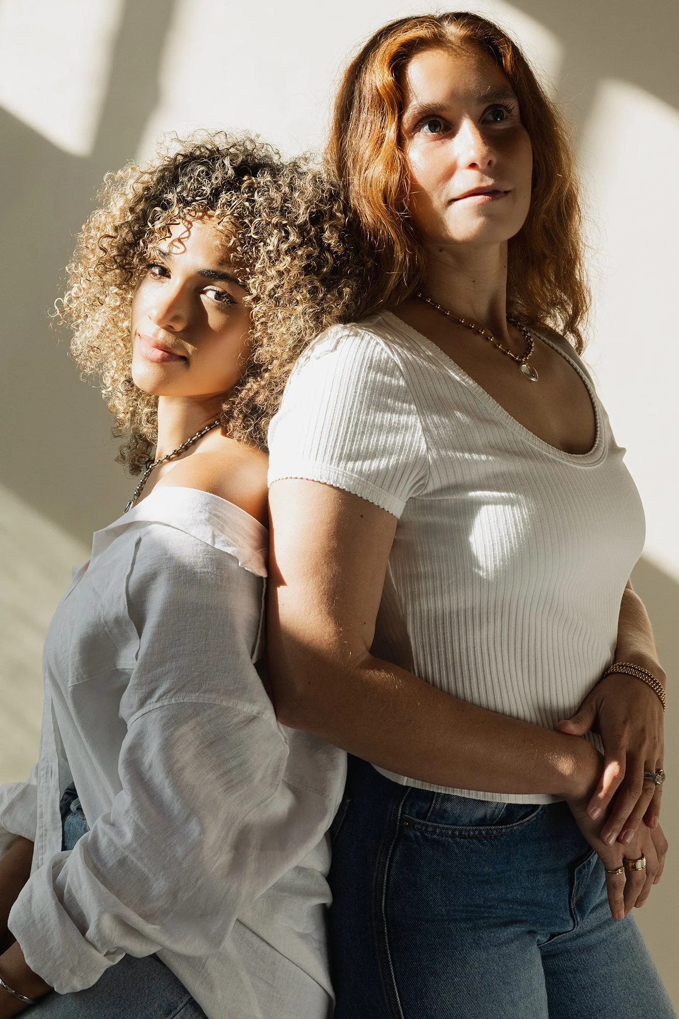 Two women standing back to back in casual clothing, both with curly hair, one with red hair and the other with light brown hair, in a well-lit room with shadows on the wall.