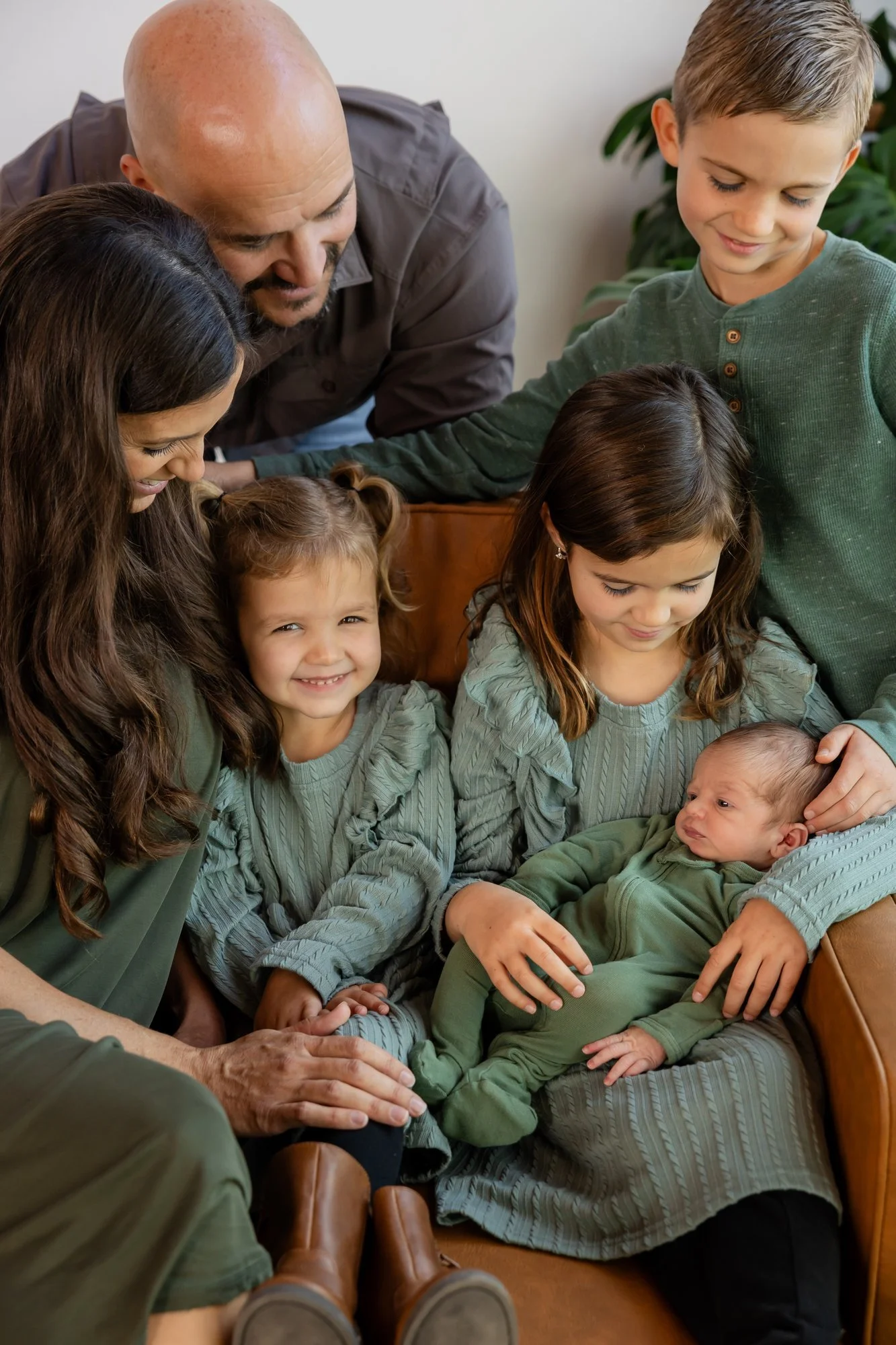 A family gathered around a newborn baby, who is lying on the woman's lap. The family members are smiling and looking at the baby, with some children touching or holding the baby. Everyone is dressed in shades of green and gray, sitting on a brown leather couch, with a plant visible in the background.