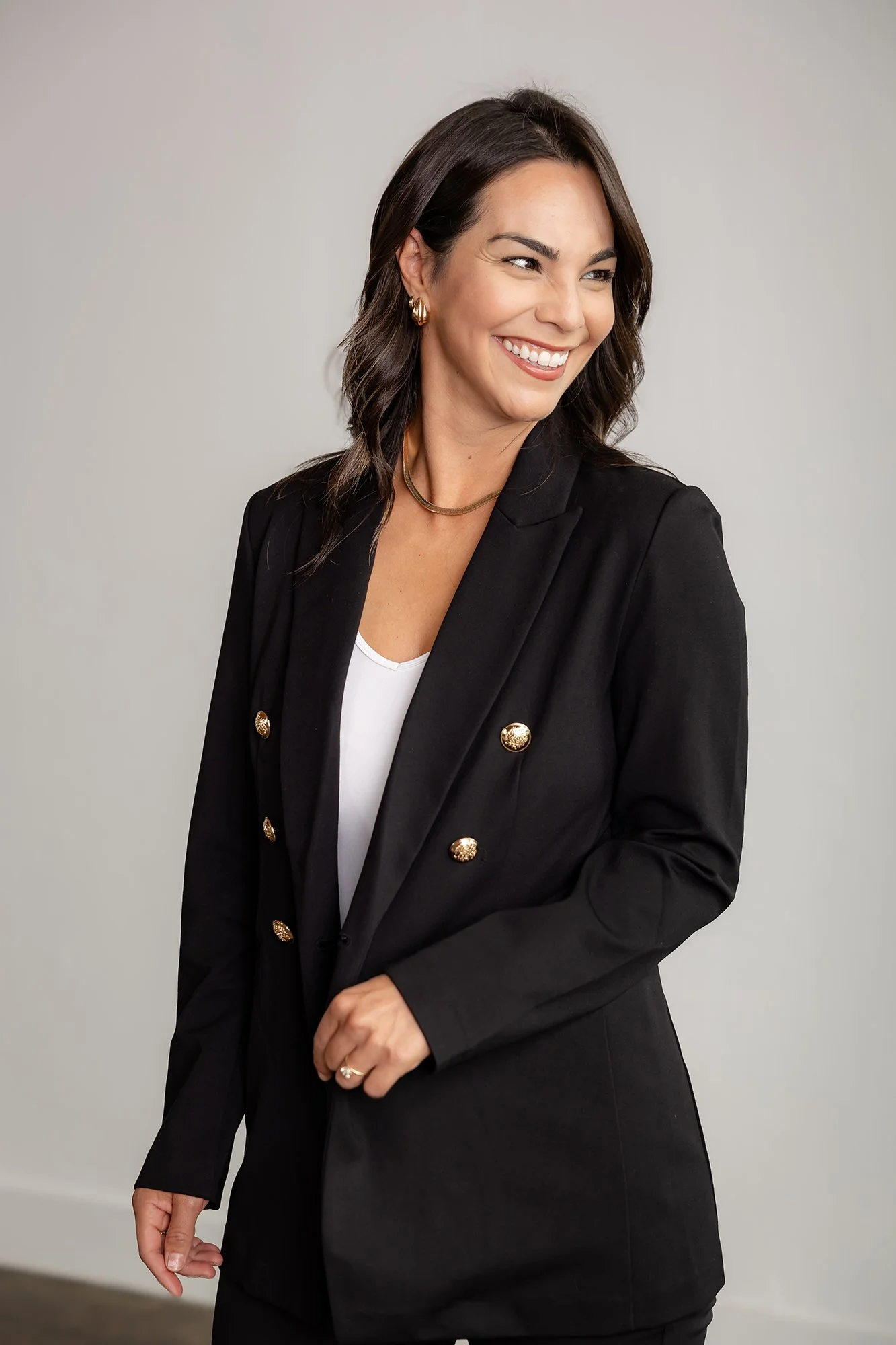 A woman with dark brown hair, styled in loose waves, smiling and looking to her left. She is wearing a black blazer with gold buttons, a white top, gold hoop earrings, and a gold necklace. Her left hand is resting near her stomach.