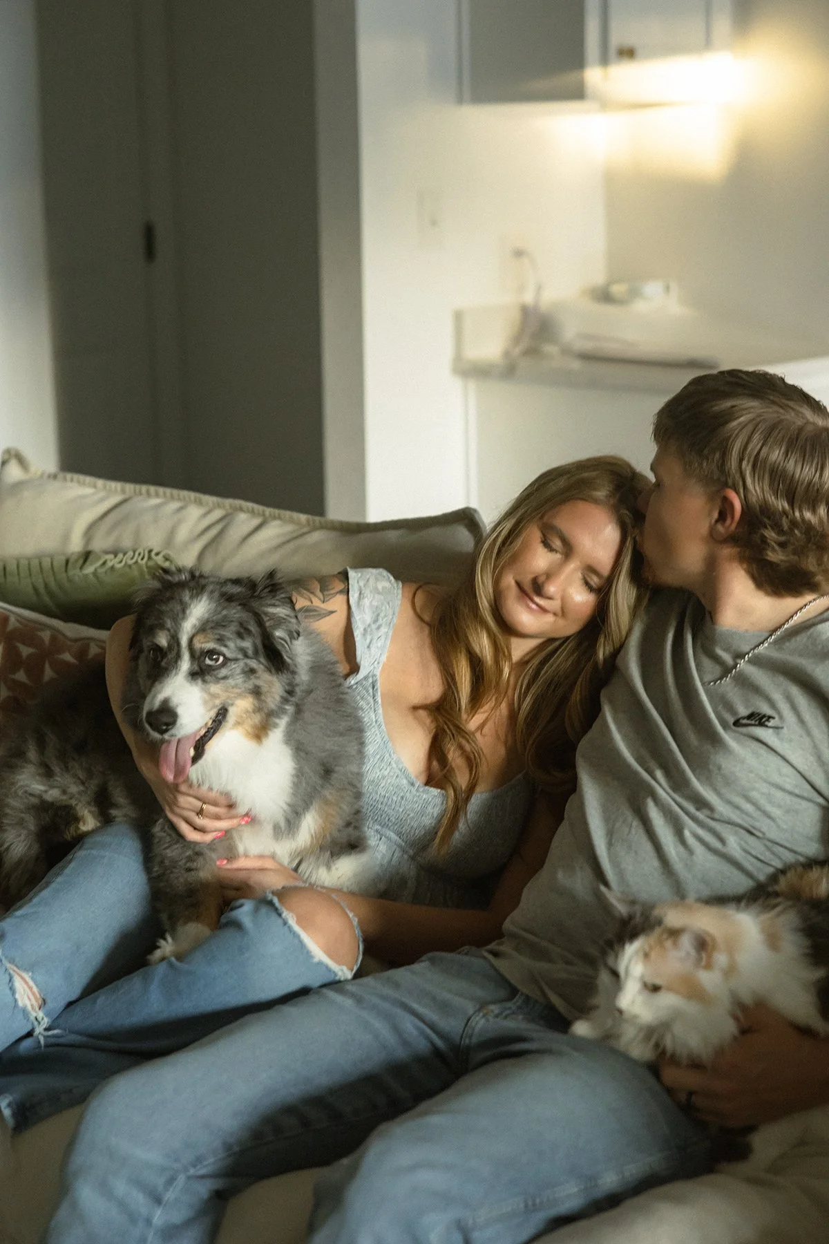 A woman and a man sitting on a couch with two dogs in a cozy home setting. The woman is holding a black and white Australian Shepherd, and the man has a small dog, possibly a Husky puppy, resting nearby.