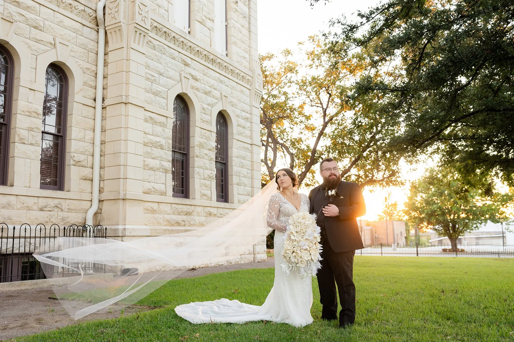 Bride and groom posing with the bride’s flowing veil outside a historic stone building at sunset in Central Texas.