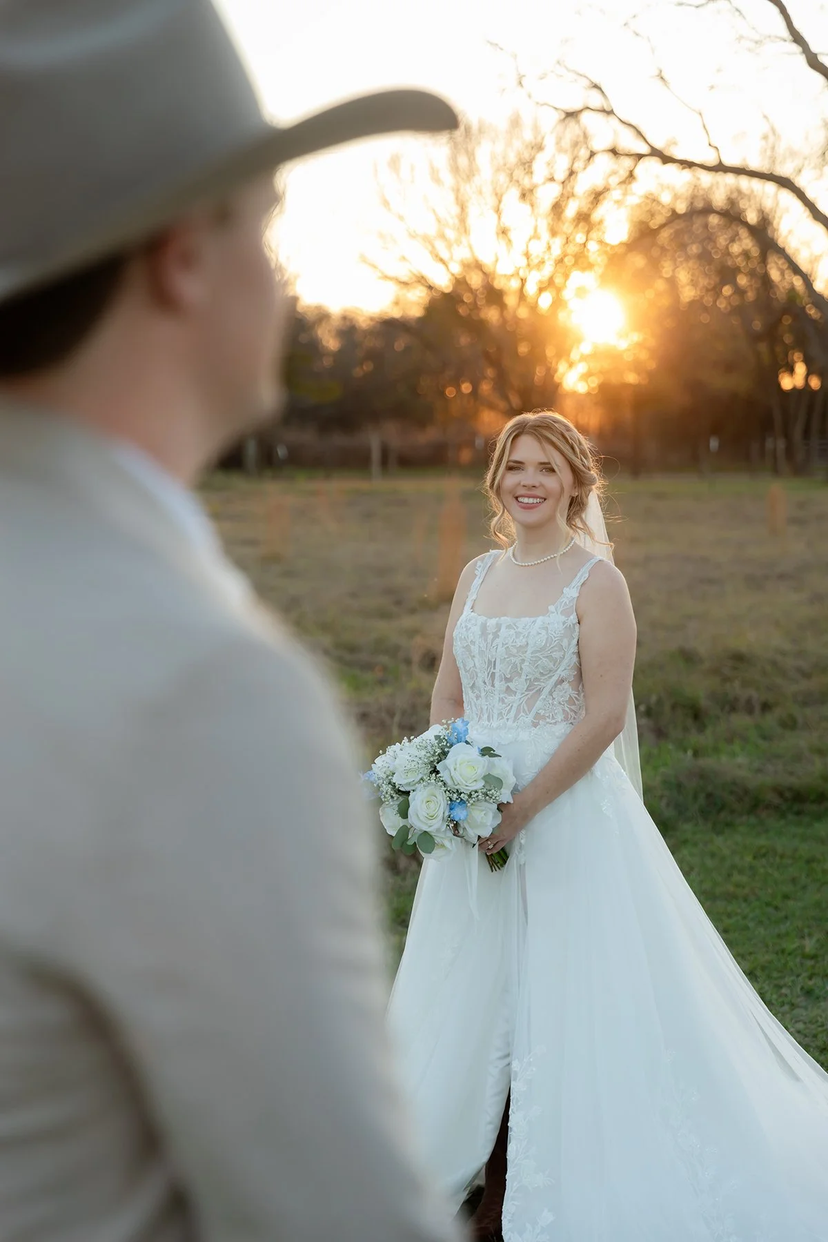 A smiling bride holding a bouquet of white and blue flowers in an outdoor setting during sunset, with trees and a field in the background.