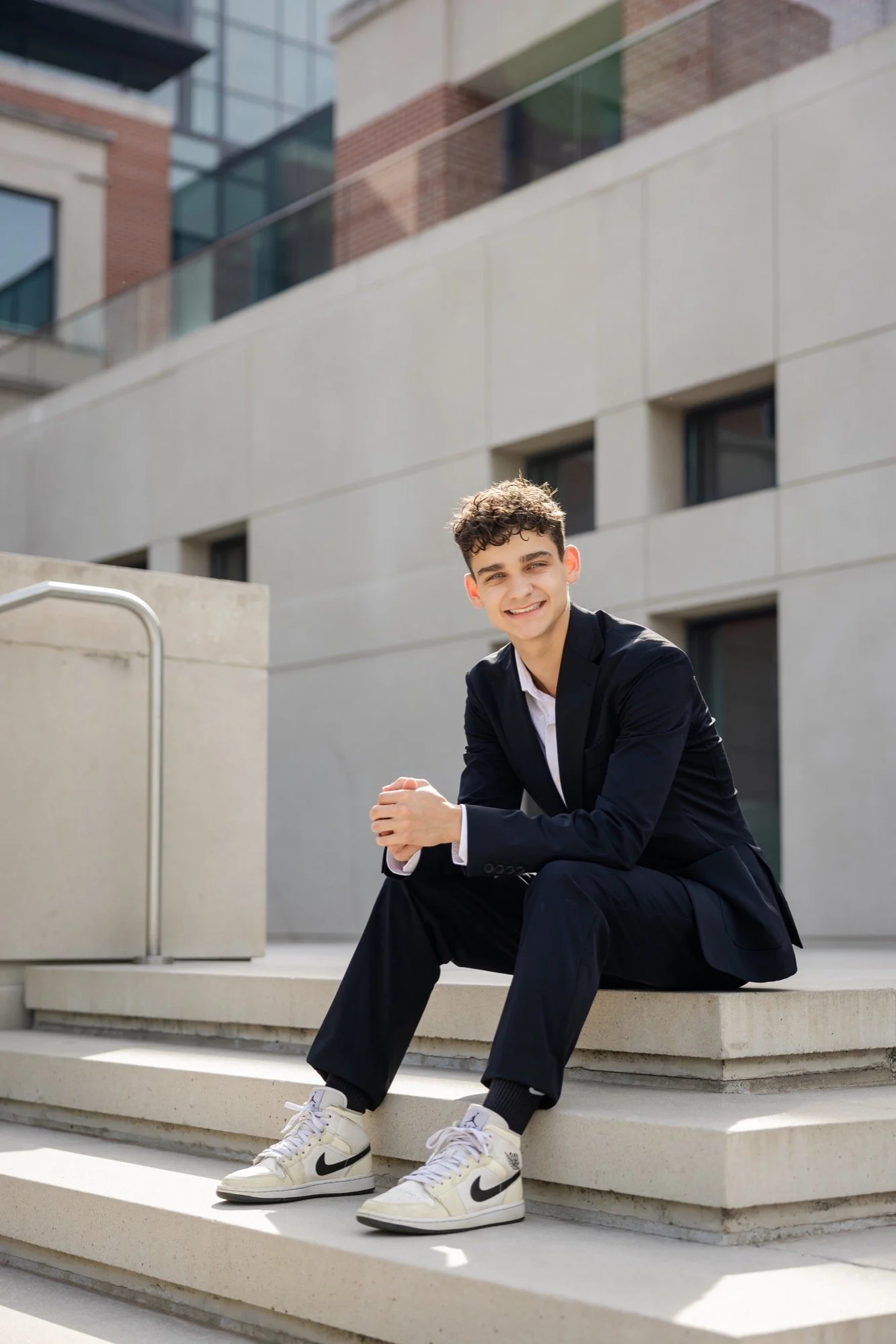 A young man with curly hair wearing a black suit, white shirt, and white Nike sneakers sitting on concrete steps outdoors, smiling at the camera in front of a modern building.