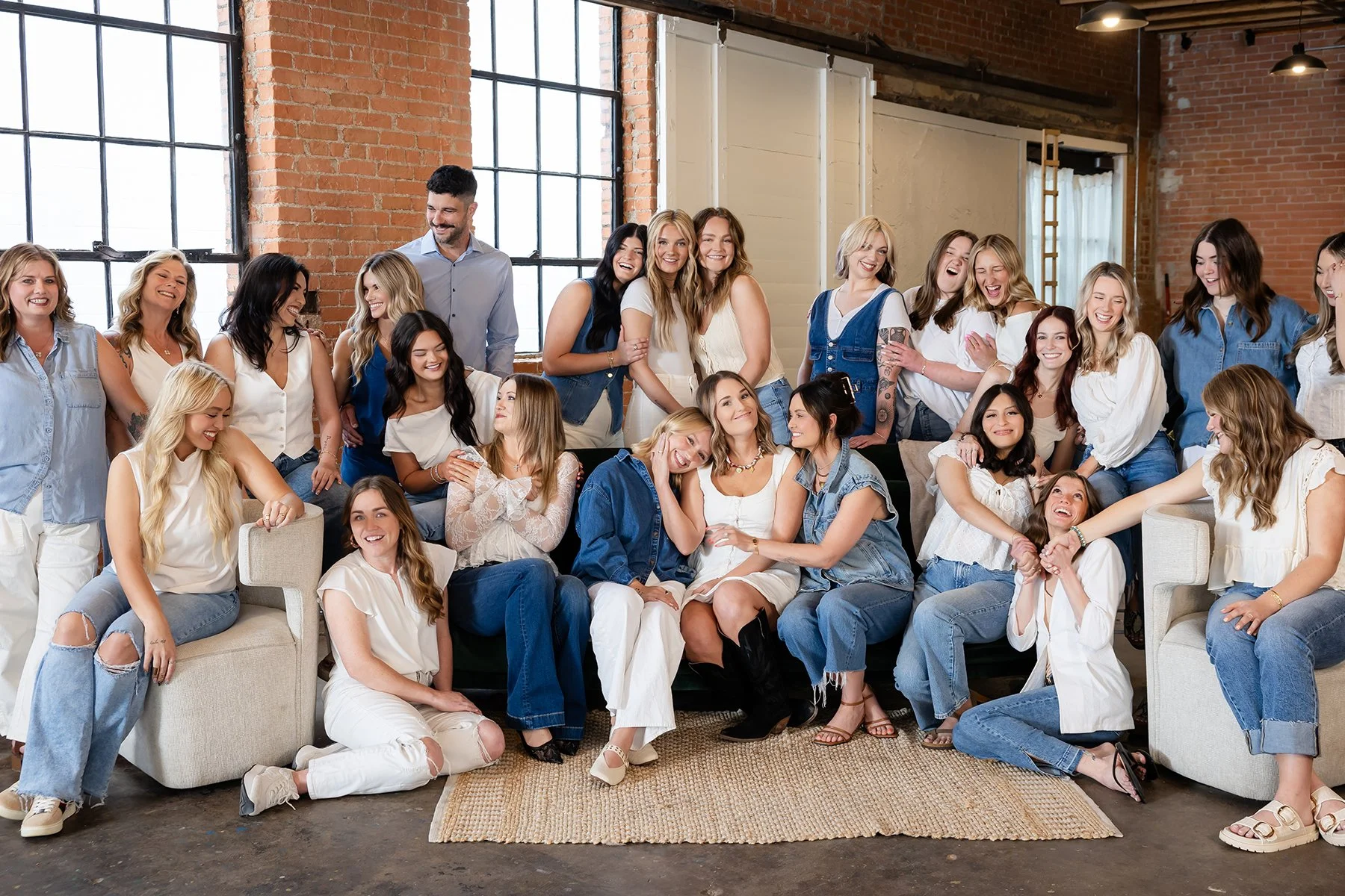 A large group of women and a man, at Sunroom Studio, sitting and standing in a cozy industrial-style area with exposed brick walls and large windows, all smiling and laughing.