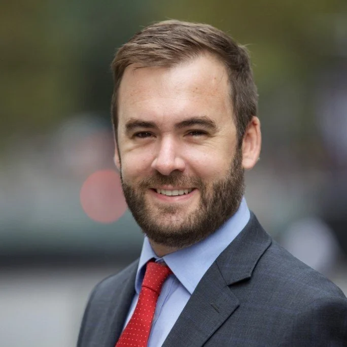 A portrait of a smiling man in a business suit, outdoors with a blurred background.