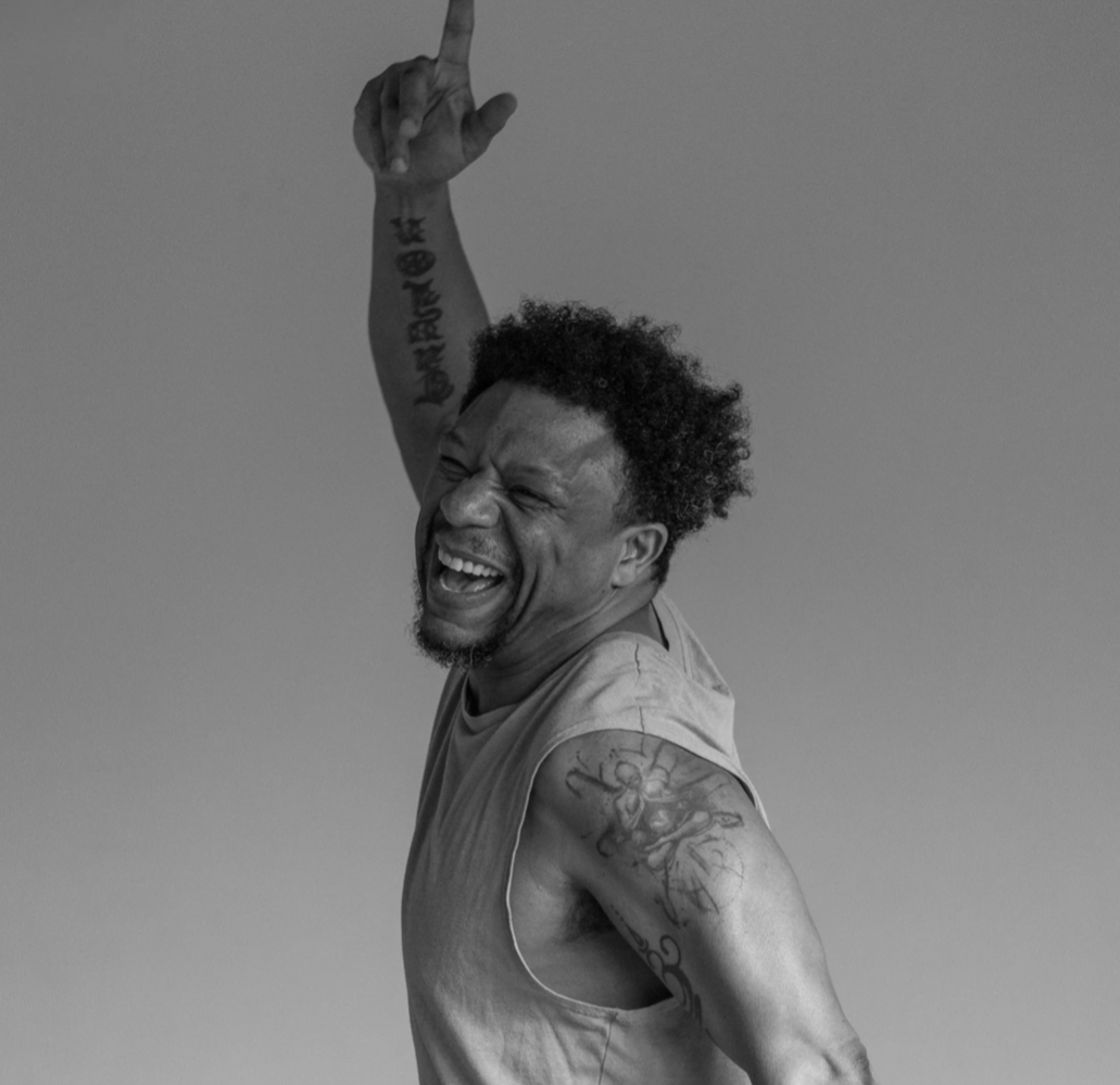 A man with tattoos on his arm and face, smiling and laughing, raising one arm with his index finger pointing upwards, against a plain background.