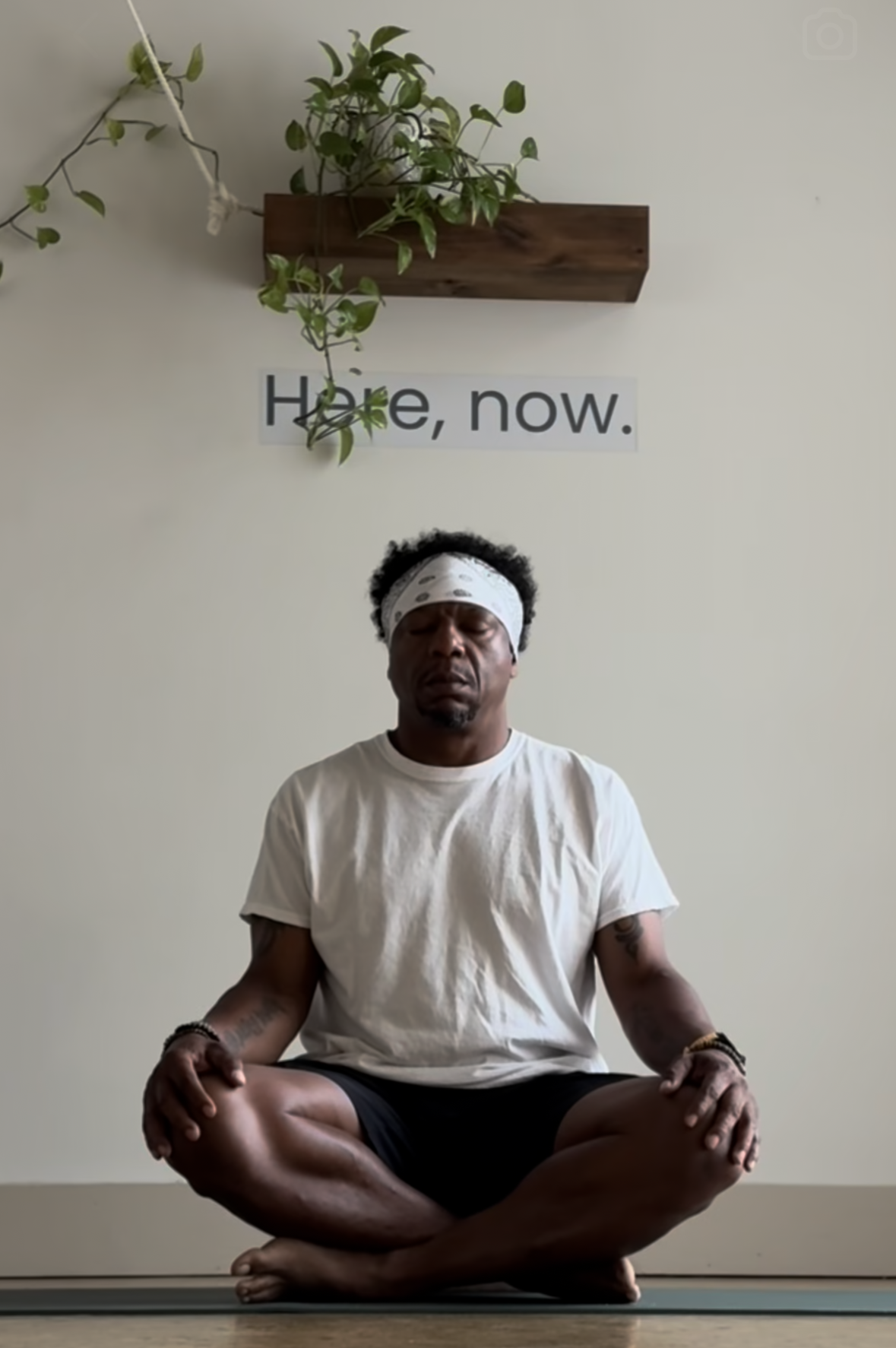 Man with a white bandana doing yoga, seated in a cross-legged pose, with a plant and the words 'Hope, now.' on the wall behind him.