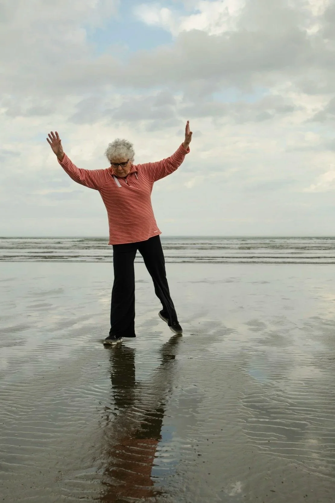 An elderly woman with curly gray hair and glasses, wearing a red and white striped long-sleeve shirt and black pants, standing on a wet beach with arms raised and reflected in the shallow water, under cloudy skies.
