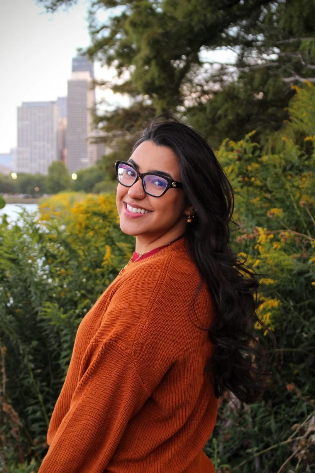 A young woman with glasses, long dark hair, and a nose ring smiling outdoors near greenery and city buildings.