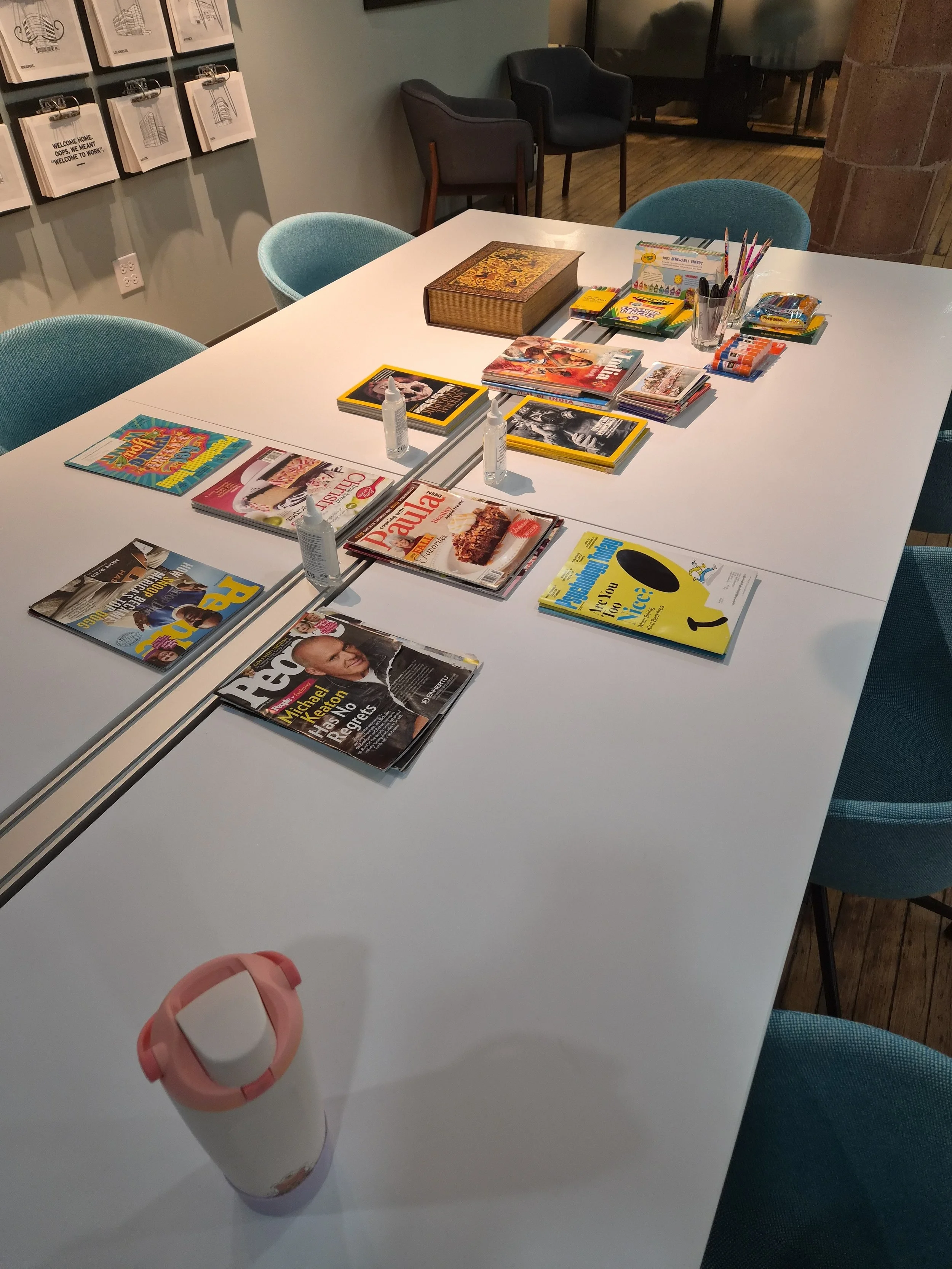 A white table with magazines, books, art supplies, and hand sanitizer bottles on it, situated in a room with chairs and framed artwork on the walls.