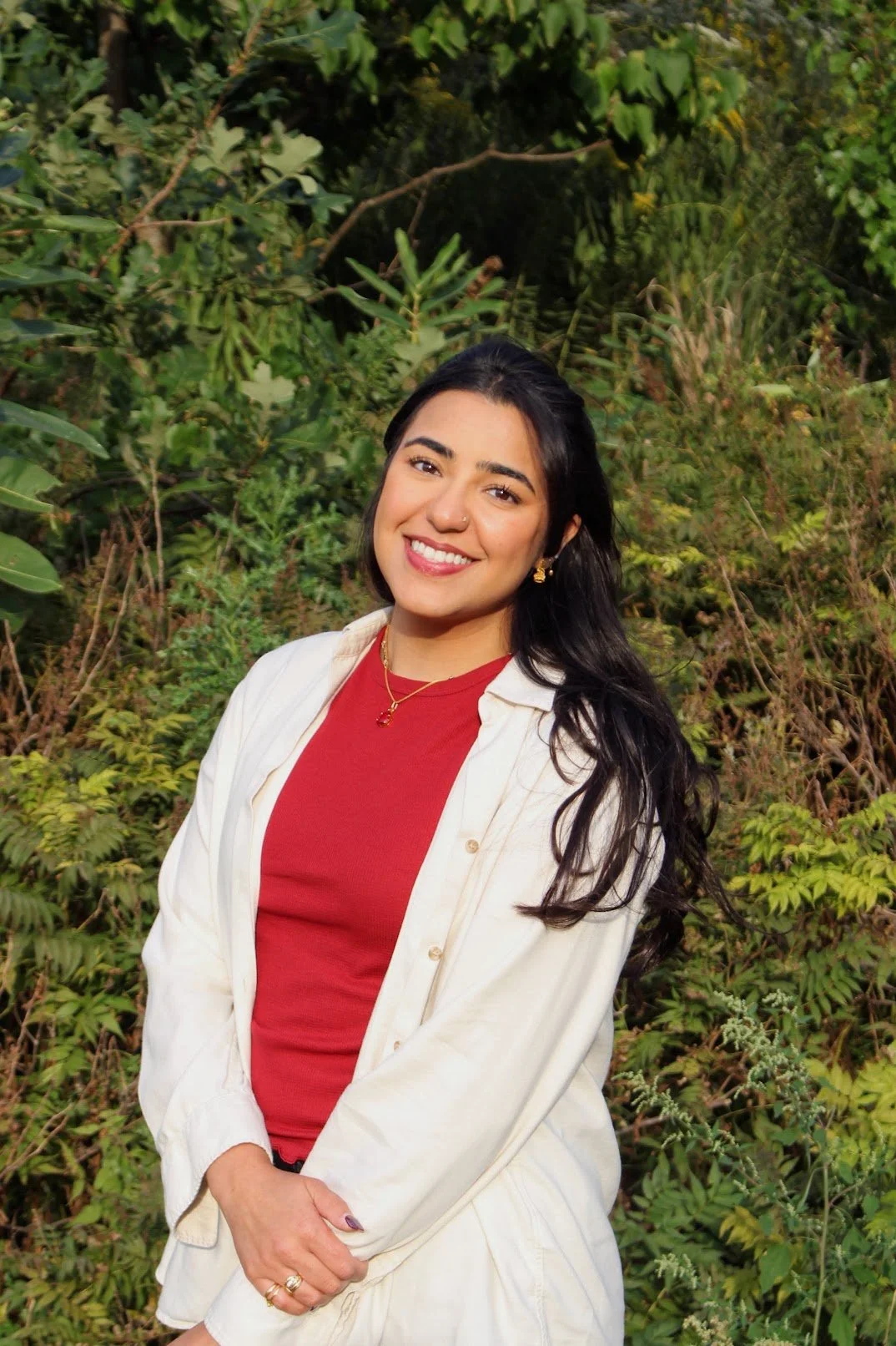 A young woman with long black hair, wearing a red top and a cream-colored jacket, smiling in front of green foliage.